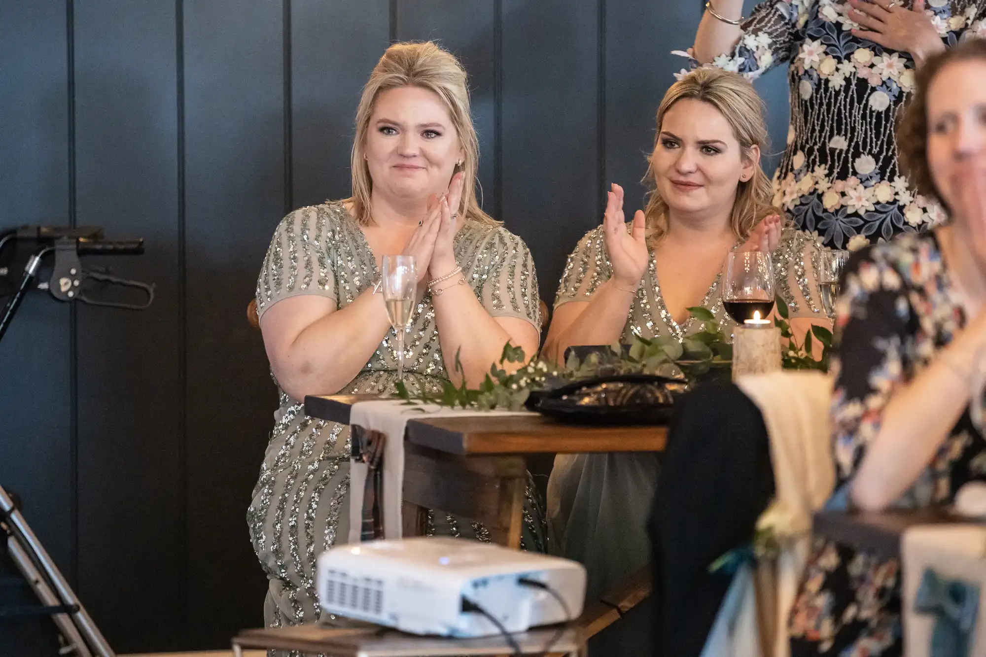 Two women in matching attire sit at a wooden table, clapping. A projector and a lit candle are on the table, and guests surround them in a decorated room.