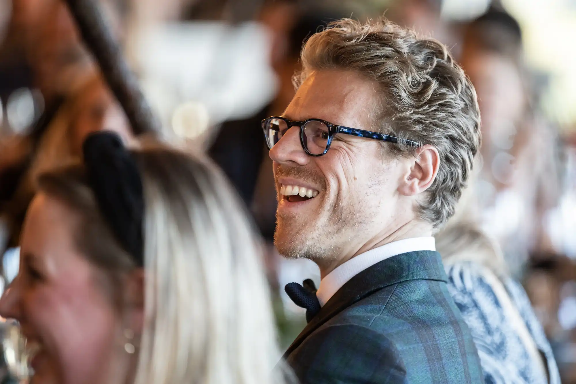 A man with glasses and a suit smiles joyfully in a crowded indoor setting.