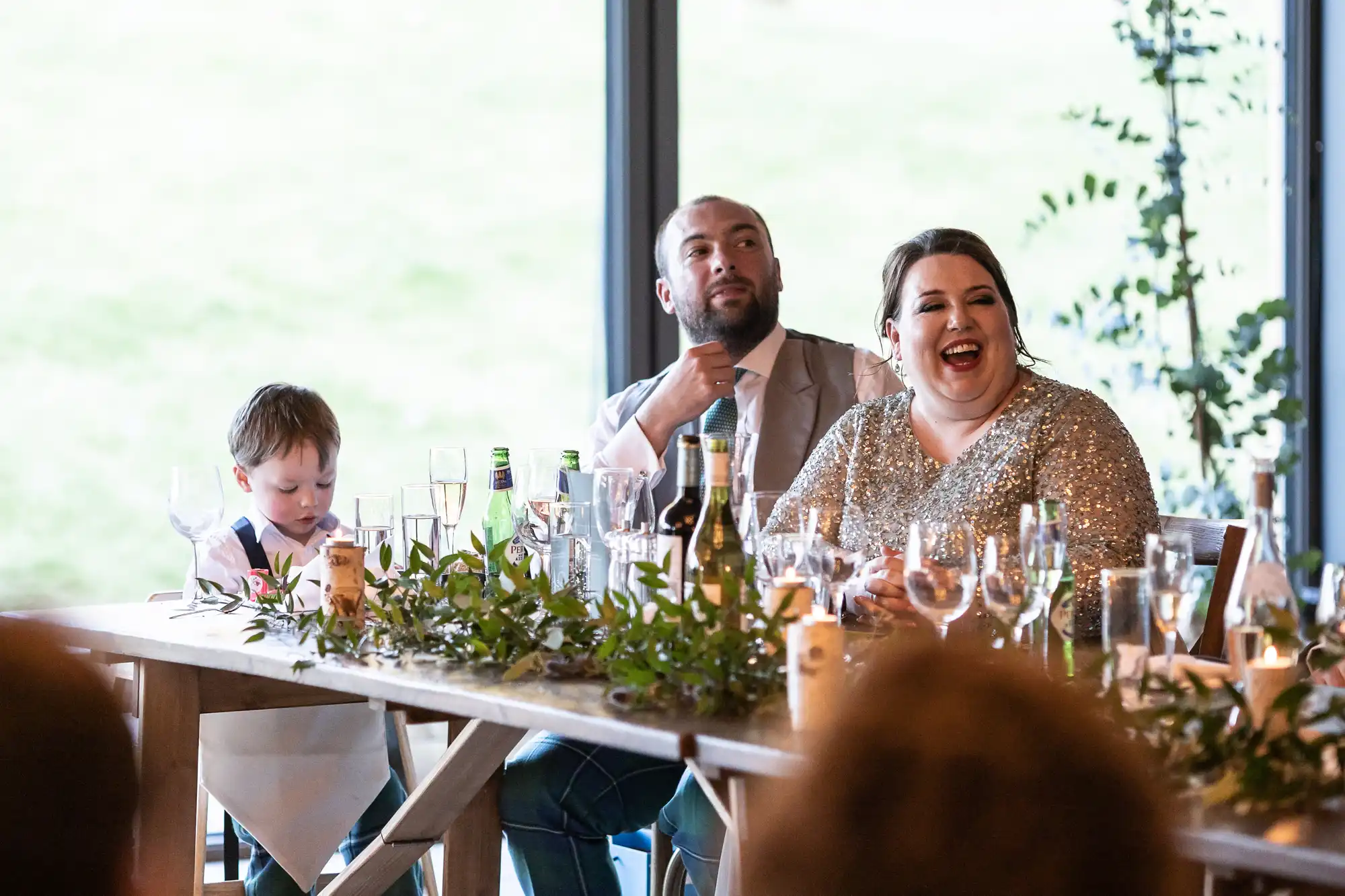 A group of people seated at a long table with food and drinks, with a woman in a sparkling dress laughing and a child sitting beside her.