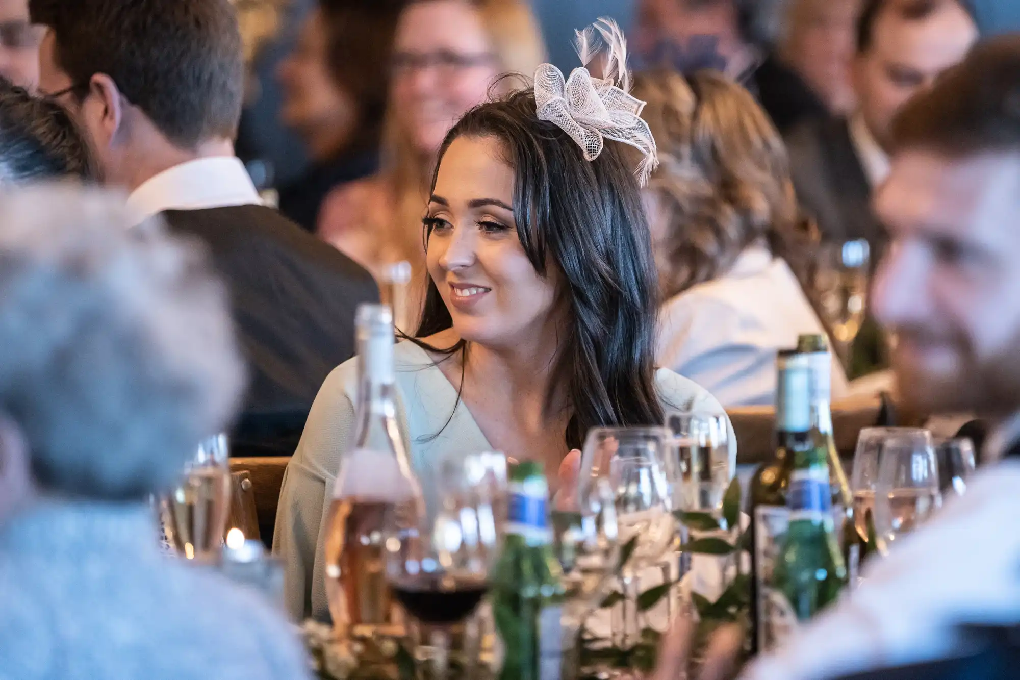 A woman with dark hair wearing a fascinator sits at a table during an event, surrounded by attendees, wine glasses, and bottles.