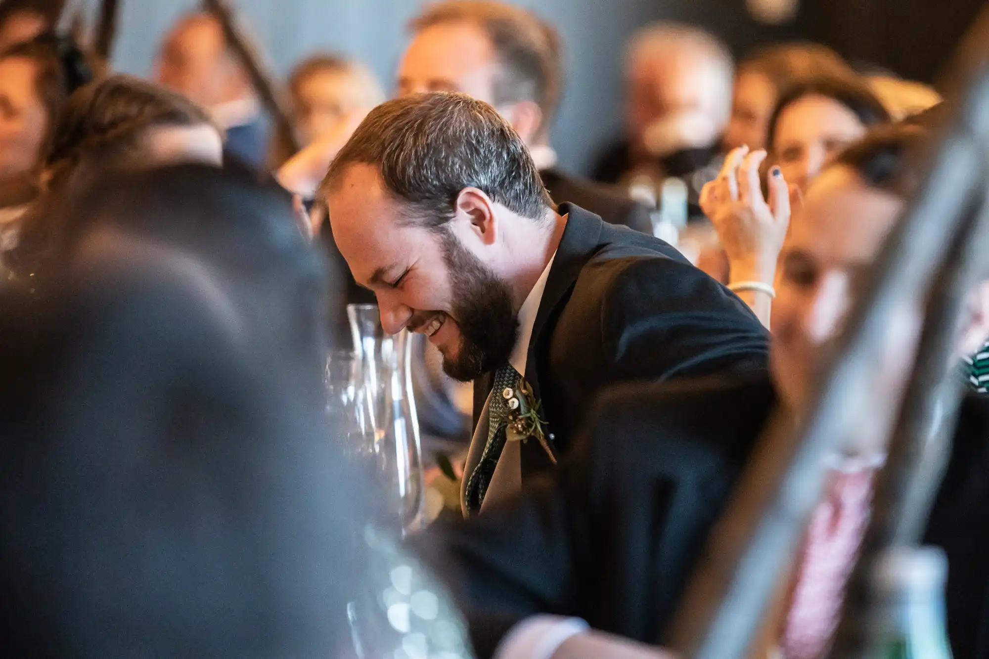 A man in a suit, sitting at a crowded event, laughs heartily with his head tilted downward.