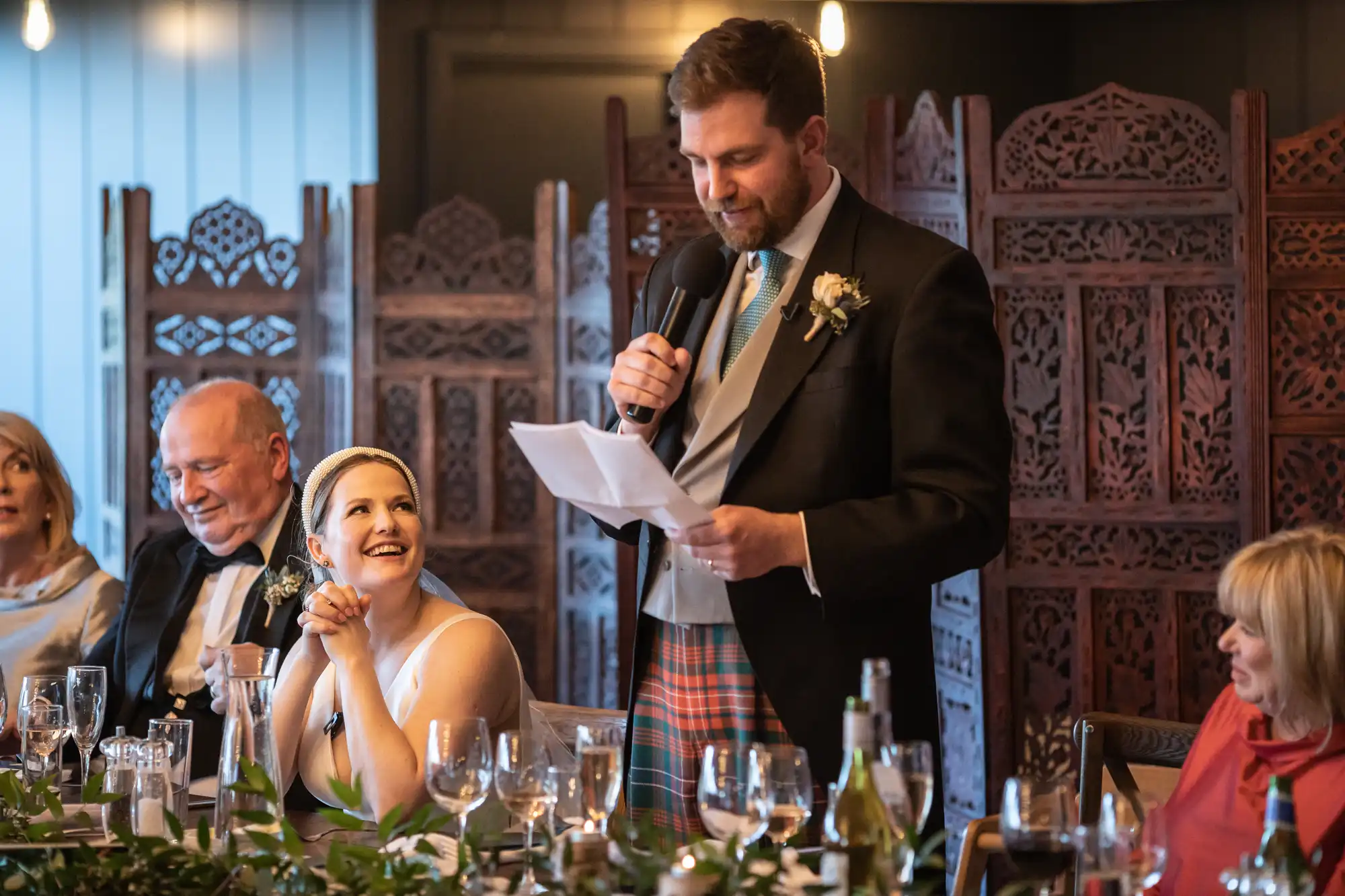 A man in formal attire speaks into a microphone while reading from a paper at a dinner event. Seated guests, including a smiling woman in a white dress, listen attentively.