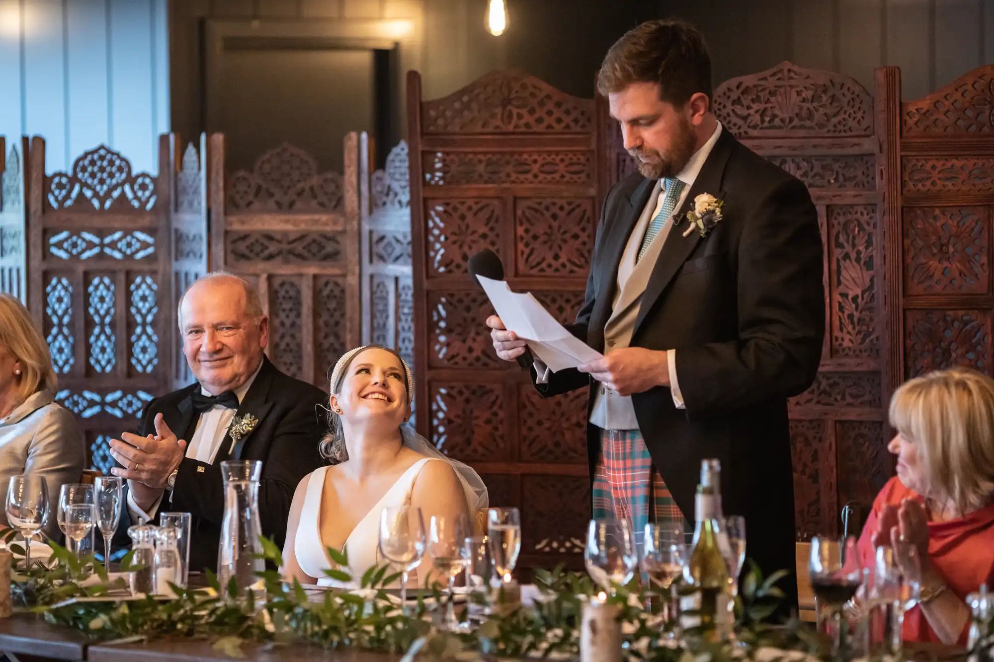 A man in formal attire gives a speech using a microphone at a wedding reception. The bride, sitting beside him, looks up smiling, while guests at the table listen.