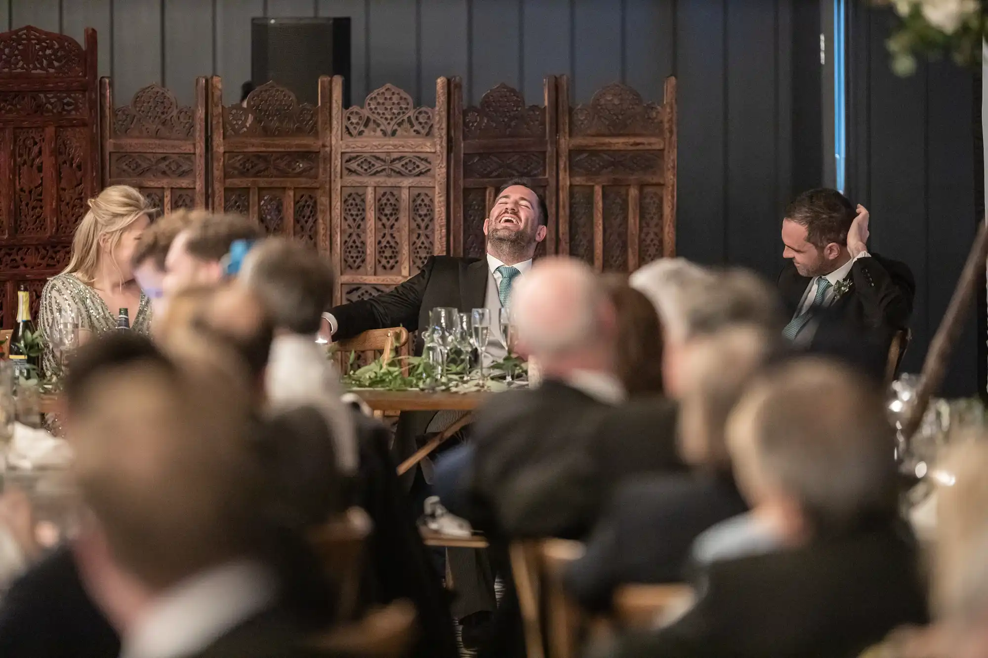 A man in a suit is laughing heartily at a formal event, while two other individuals at the table and several blurred attendees are visible in the foreground. An ornate wooden screen is in the background.