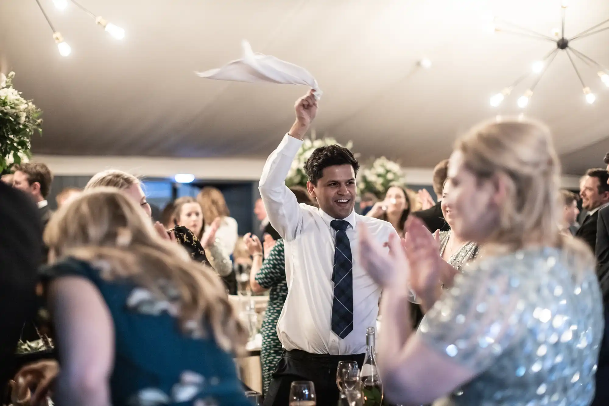 A man in a white shirt and tie waves a napkin in celebration at an indoor event surrounded by people clapping.