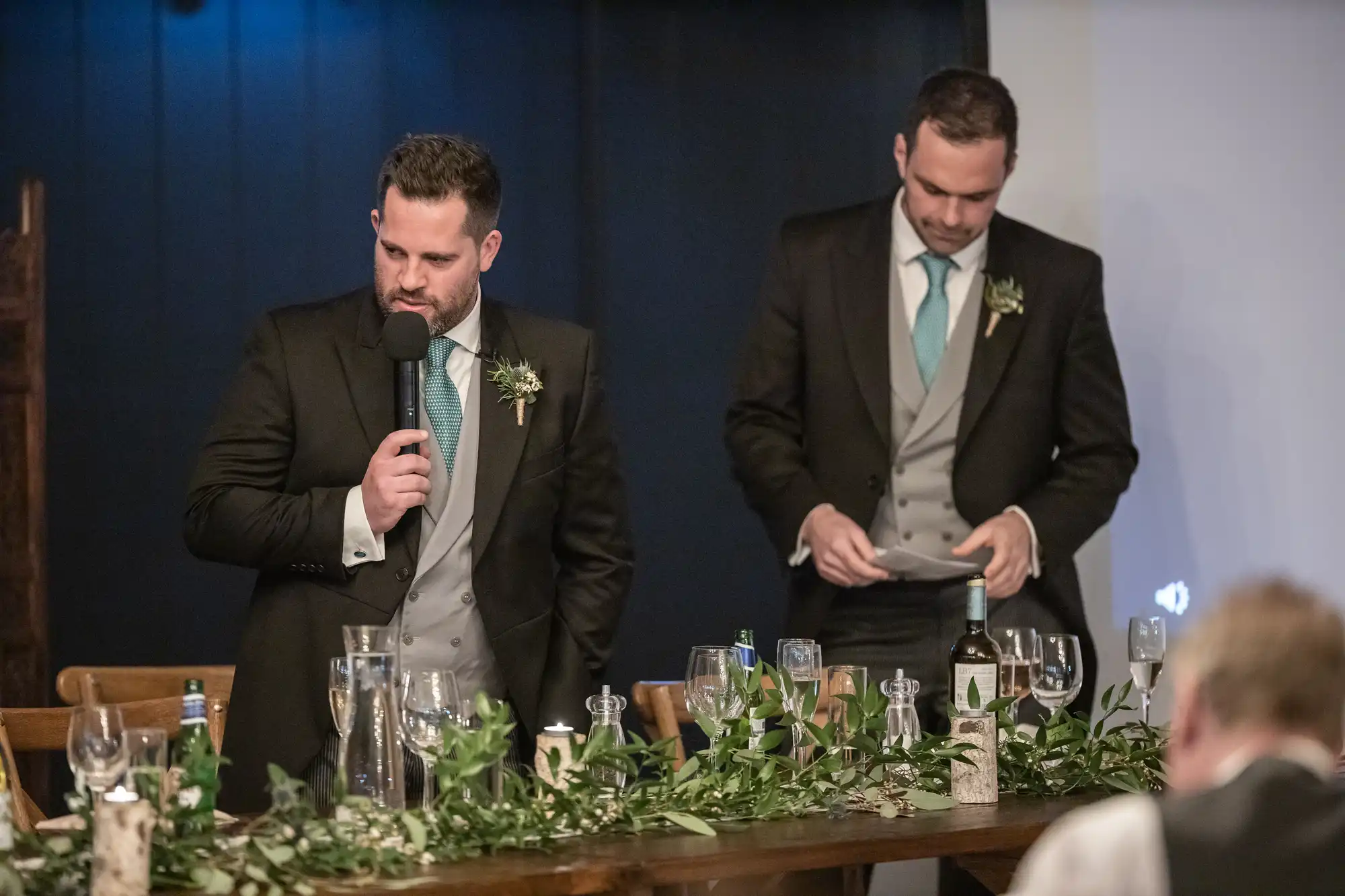 Two men in formal attire stand by a decorated table. One speaks into a microphone, while the other looks at a note. Greenery and empty glasses are on the table.