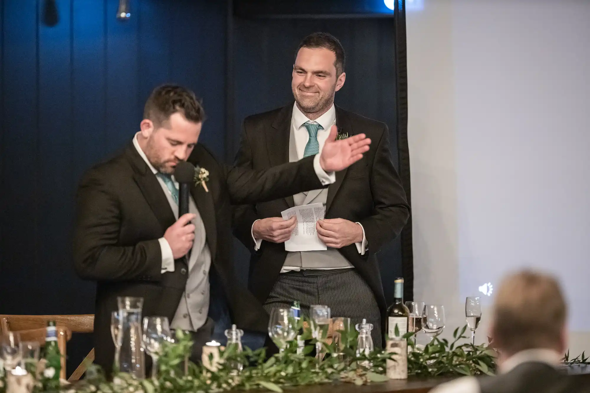 Two men in suits stand behind a table with a green table runner and wine glasses. One man is speaking into a microphone while the other looks on and holds papers.