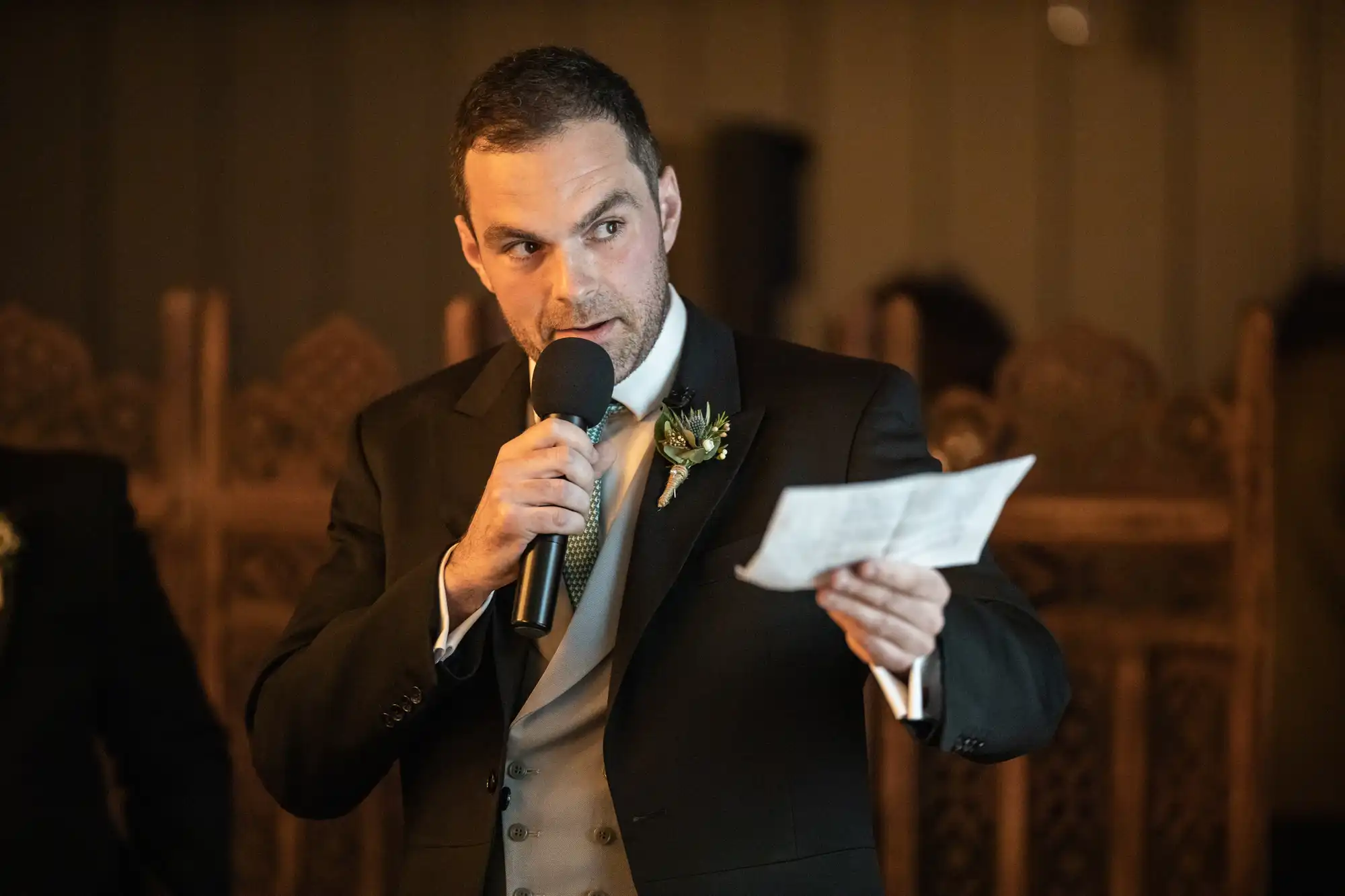 Man in a suit holding a microphone and reading from a piece of paper while speaking indoors.