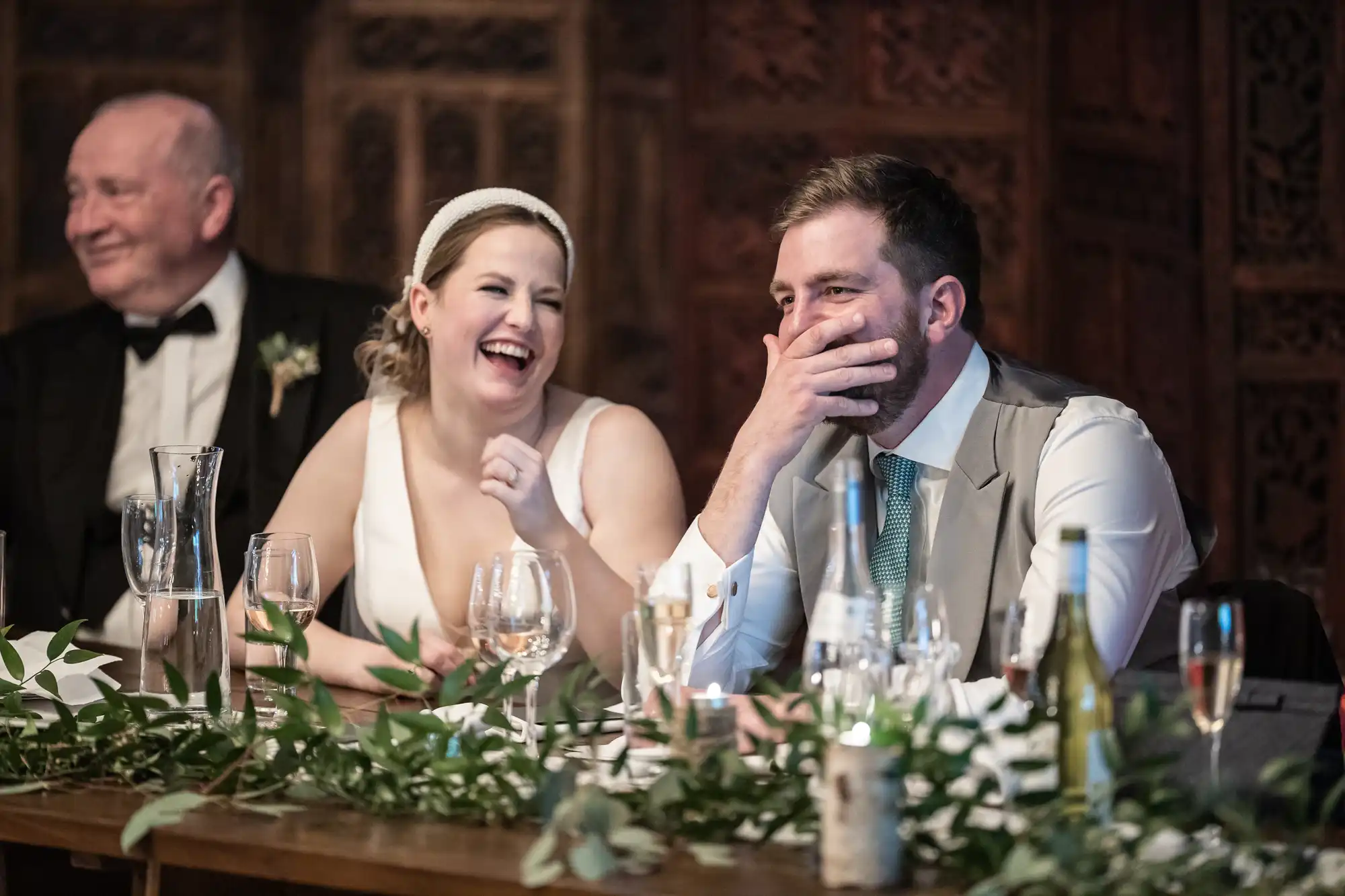 A bride and groom sit at a table, laughing together. The bride wears a white dress and headband, while the groom is in a vest and tie. A man in formal attire sits to their left. Various drinks are on the table.