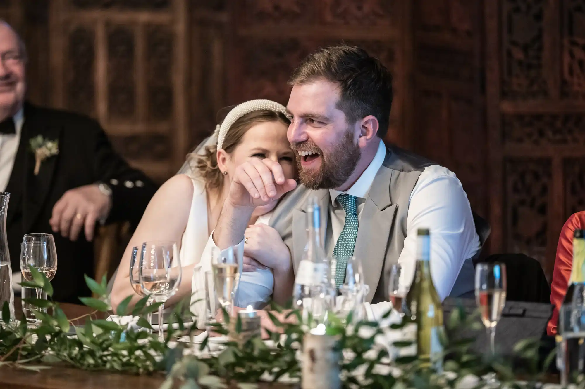 A bride and groom sit at a wedding reception table, smiling and laughing together. Guests and glasses are around them, with greenery as table decor.