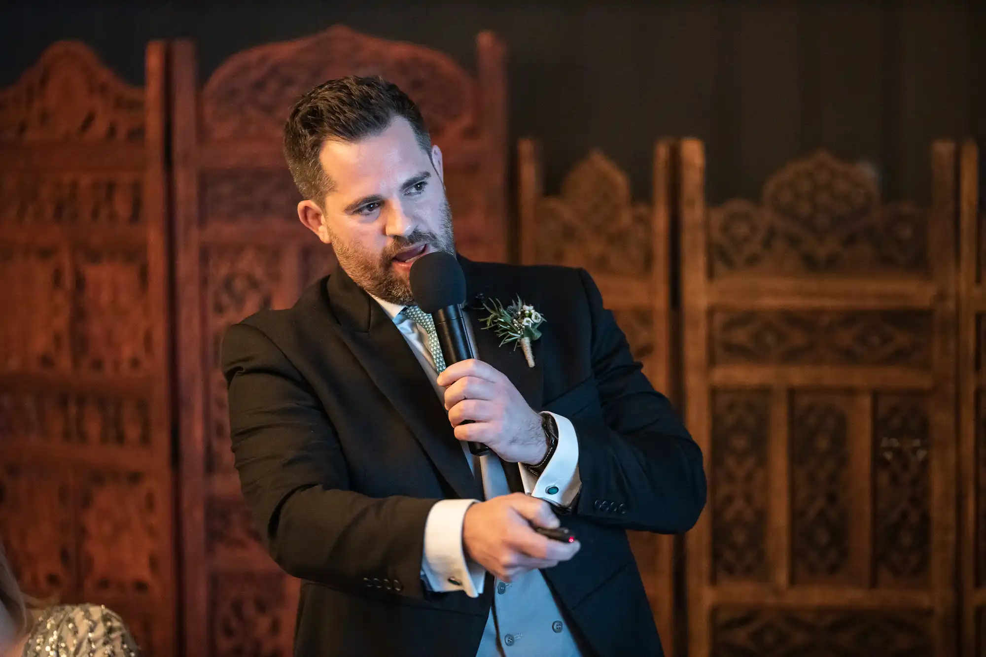 A man in a dark suit and tie is speaking into a microphone and gesturing with his hand while standing in front of ornate wooden panels.