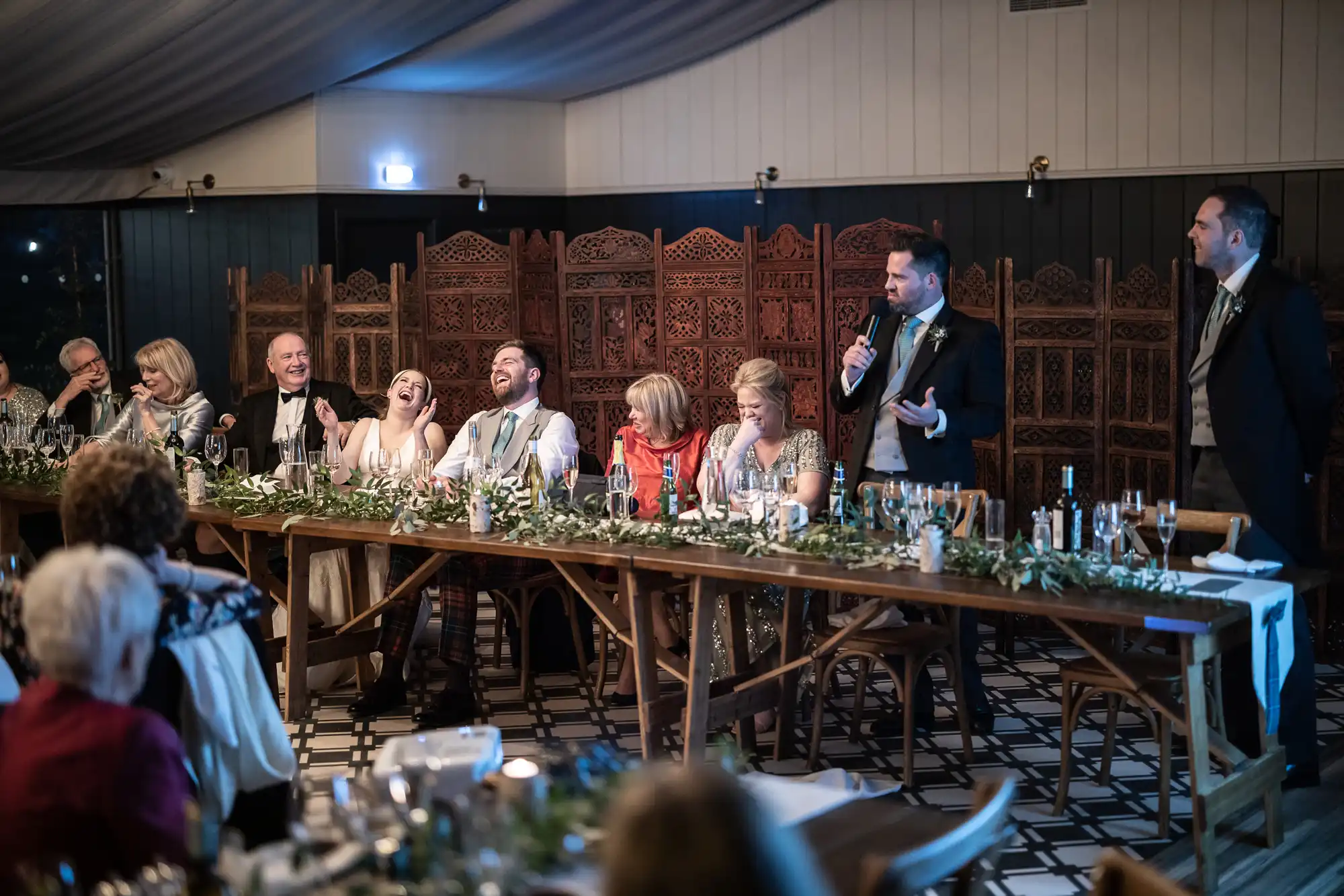 A group of people in formal attire sit at a long table with green floral arrangements, laughing and listening to a man standing and speaking into a microphone.