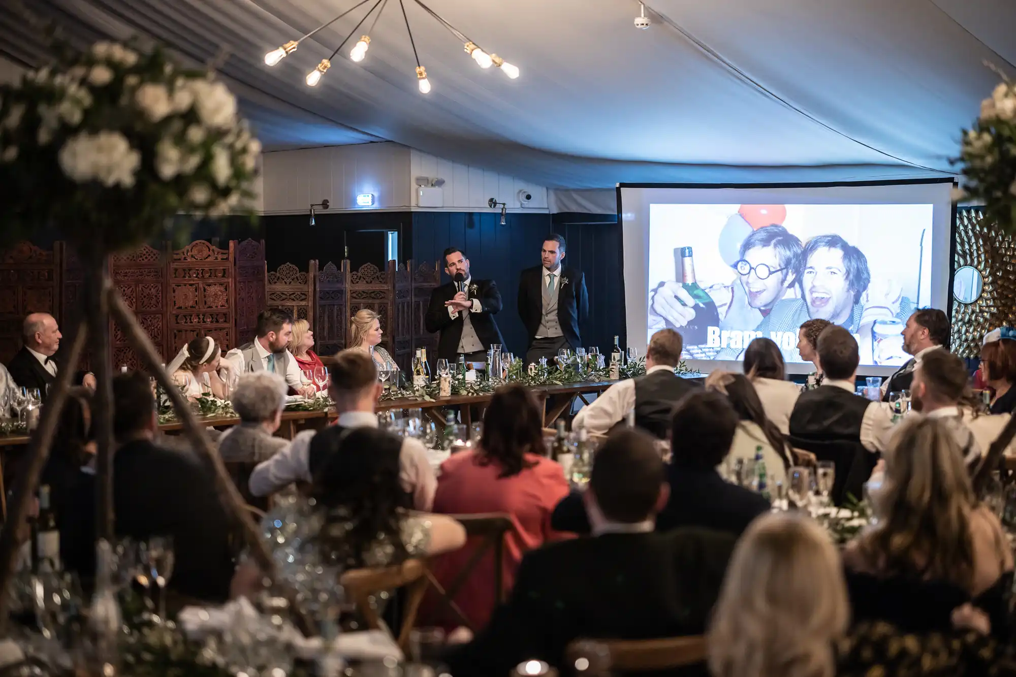 A group of people seated at tables watches two men give a speech in front of a large presentation screen at a formal event.