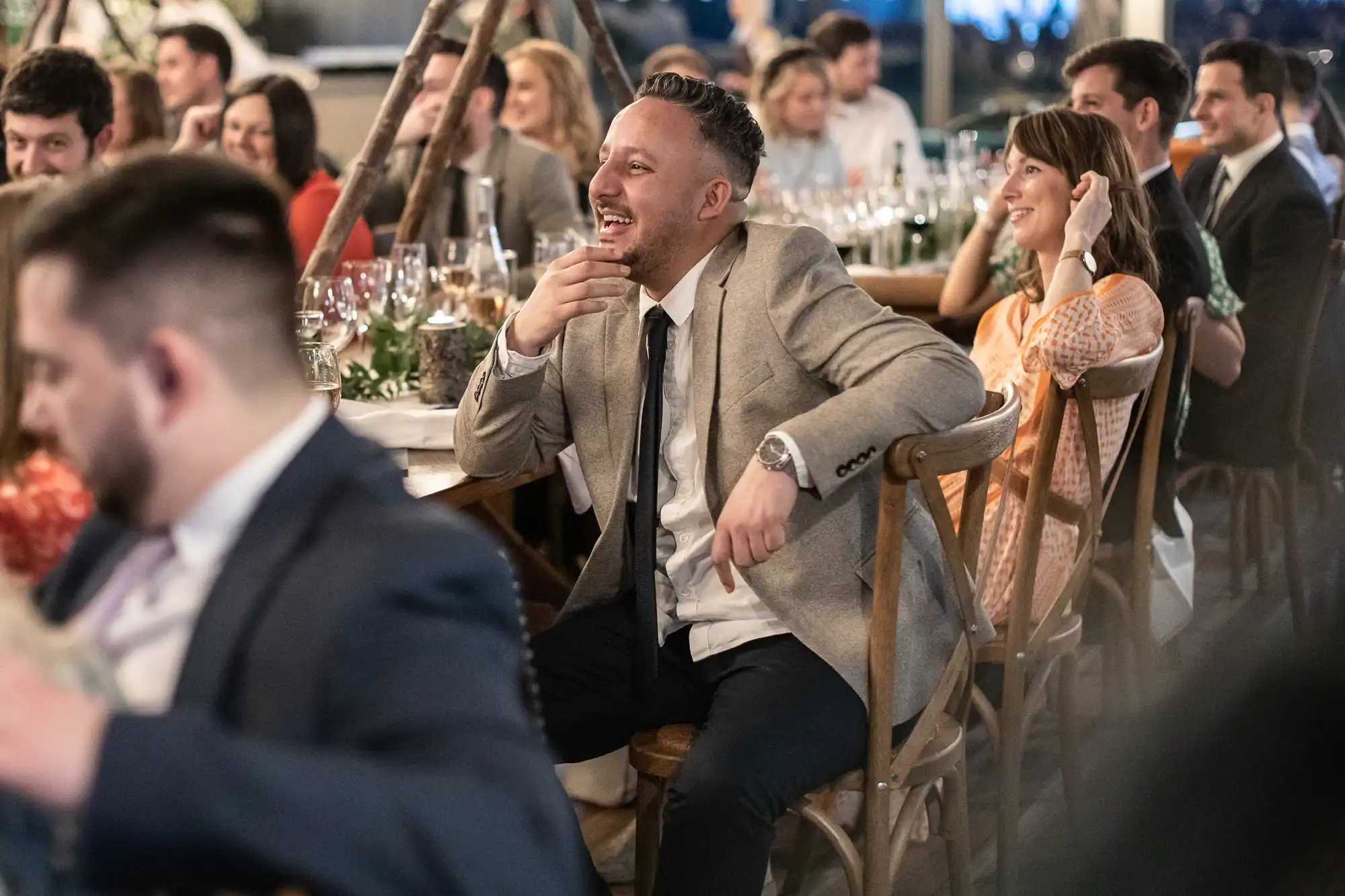 People are seated at a banquet, with a man in a gray jacket and black tie prominently laughing at the center. Other attendees are engaged and smiling. The setting appears festive and lively.