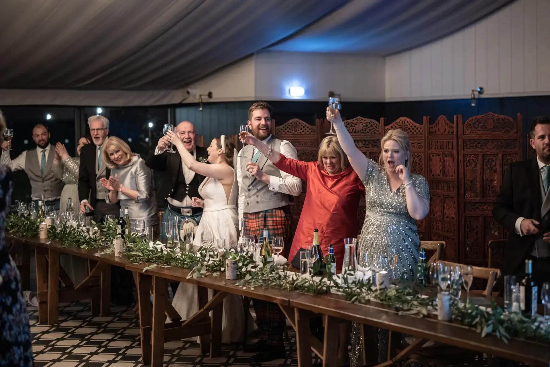 A group of people in formal attire stands and raises glasses in a toast at a wedding reception, with a long table decorated with greenery and candles in the foreground.
