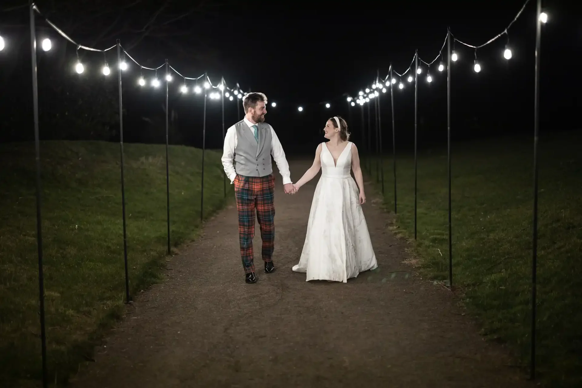 Wedding photographer Edinburgh photo of a couple holds hands while walking down an outdoor path at night, illuminated by string lights. The man wears a vest and tartan trousers, and the woman wears a white dress.