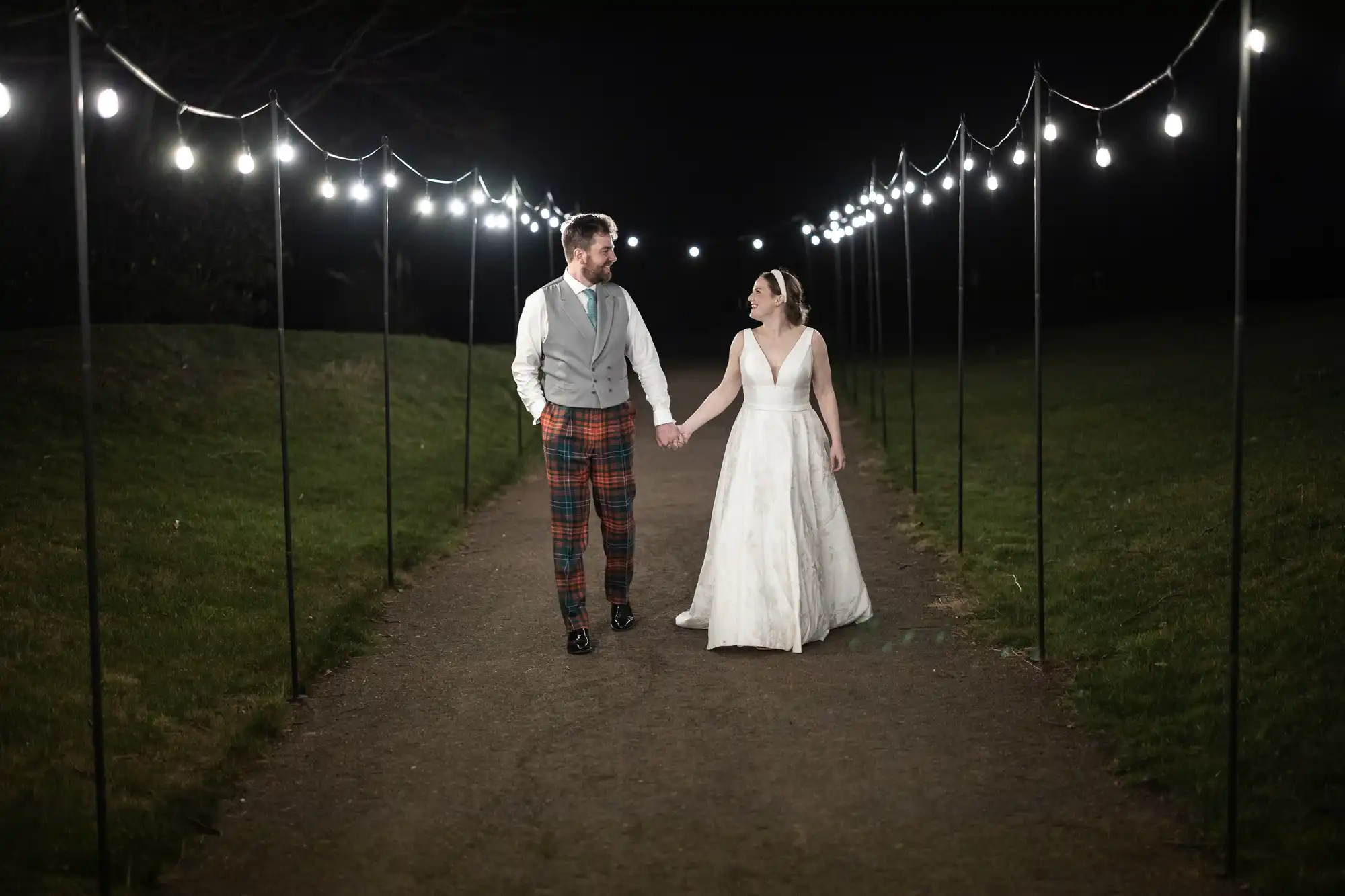 A couple holds hands while walking down an outdoor path at night, illuminated by string lights. The man wears a vest and plaid pants, and the woman wears a white dress.