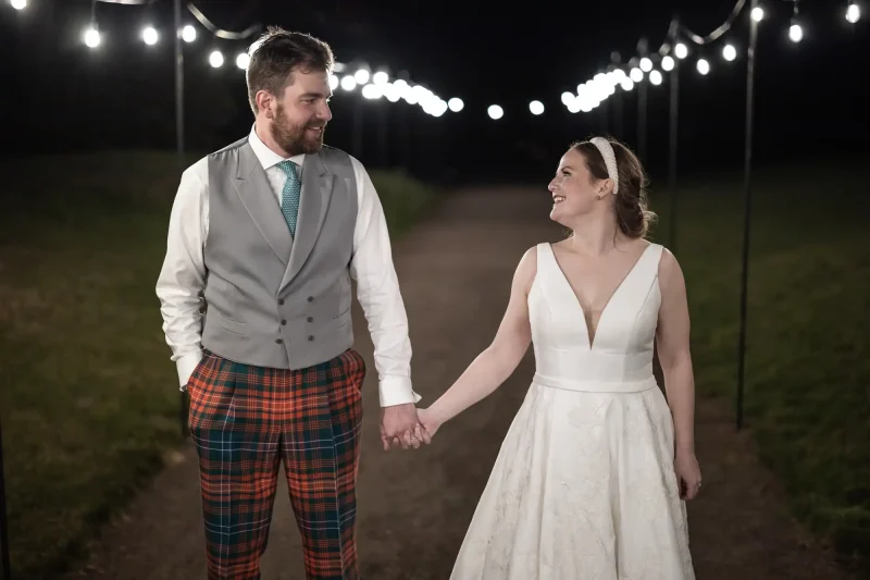 Dunglass Estate wedding photography: A couple dressed in wedding attire holds hands and smiles at each other under a string of lights, standing on a path at night.