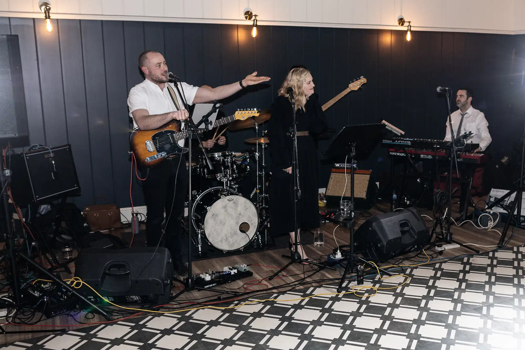 A band performing live indoors with a guitarist, drummer, vocalist, bassist, and keyboardist. Instruments and equipment are set up on a patterned floor with dark paneled walls in the background.