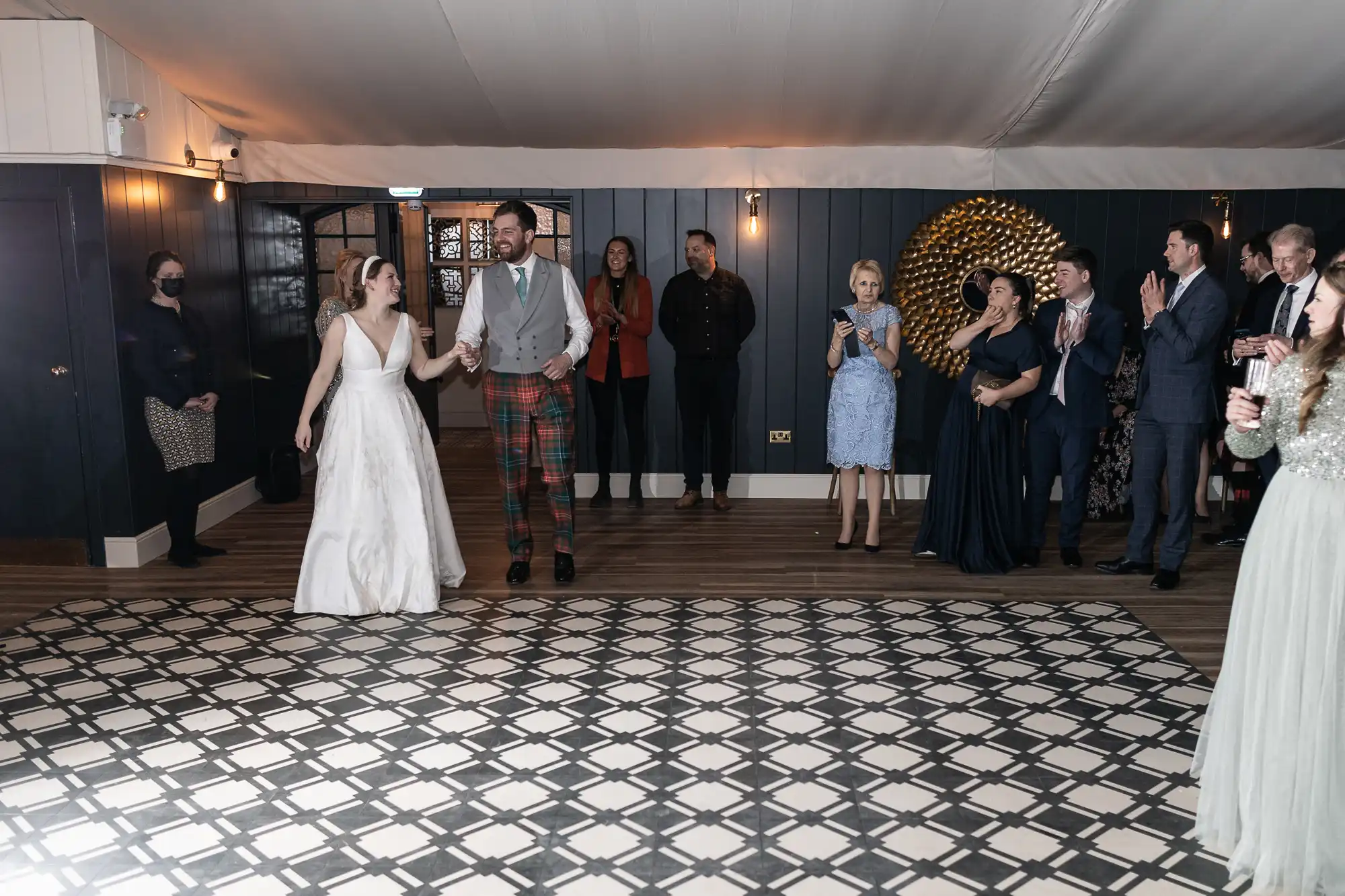 A newlywed couple holds hands and smiles while walking across a patterned floor, surrounded by friends and family who are clapping and watching them.