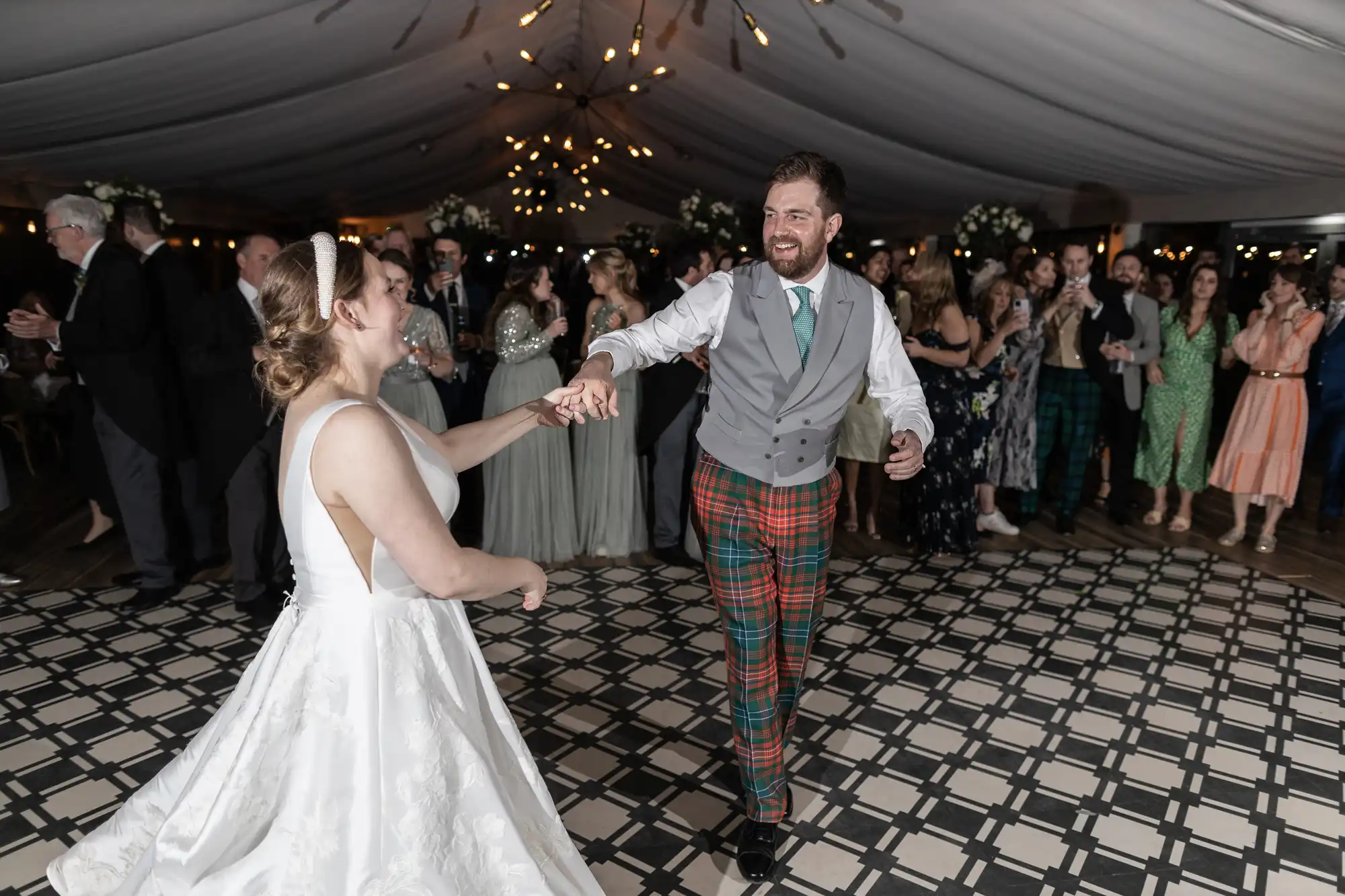 A bride and groom dance at their wedding reception, surrounded by guests. The groom wears a grey vest with plaid pants, and the bride is in a white gown. The venue is decorated with lights and flowers.