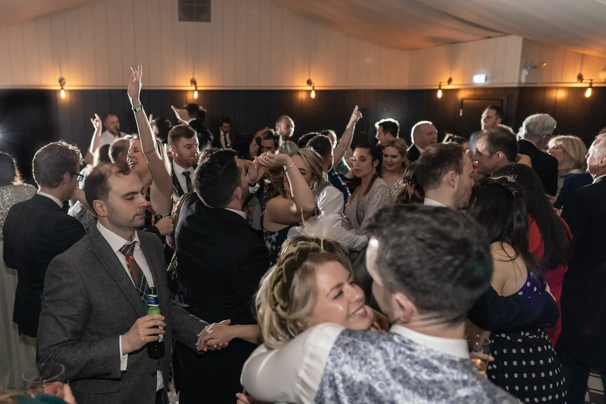 A large group of people in formal attire are socializing and dancing at an indoor event. Some are smiling and hugging, while others chat and hold drinks. The dim lighting creates a warm ambiance.