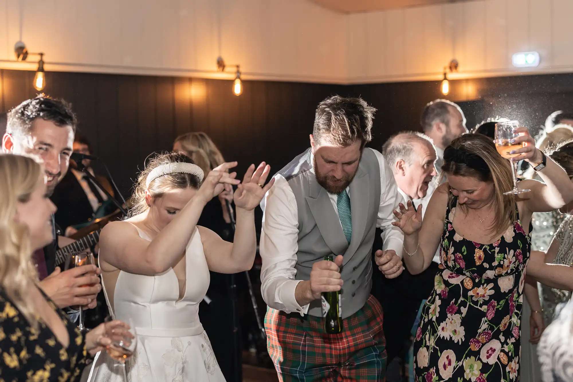 A group of people are dancing at an indoor event. The man in the center is wearing plaid pants and holding a bottle, while others around him raise their hands and smile.