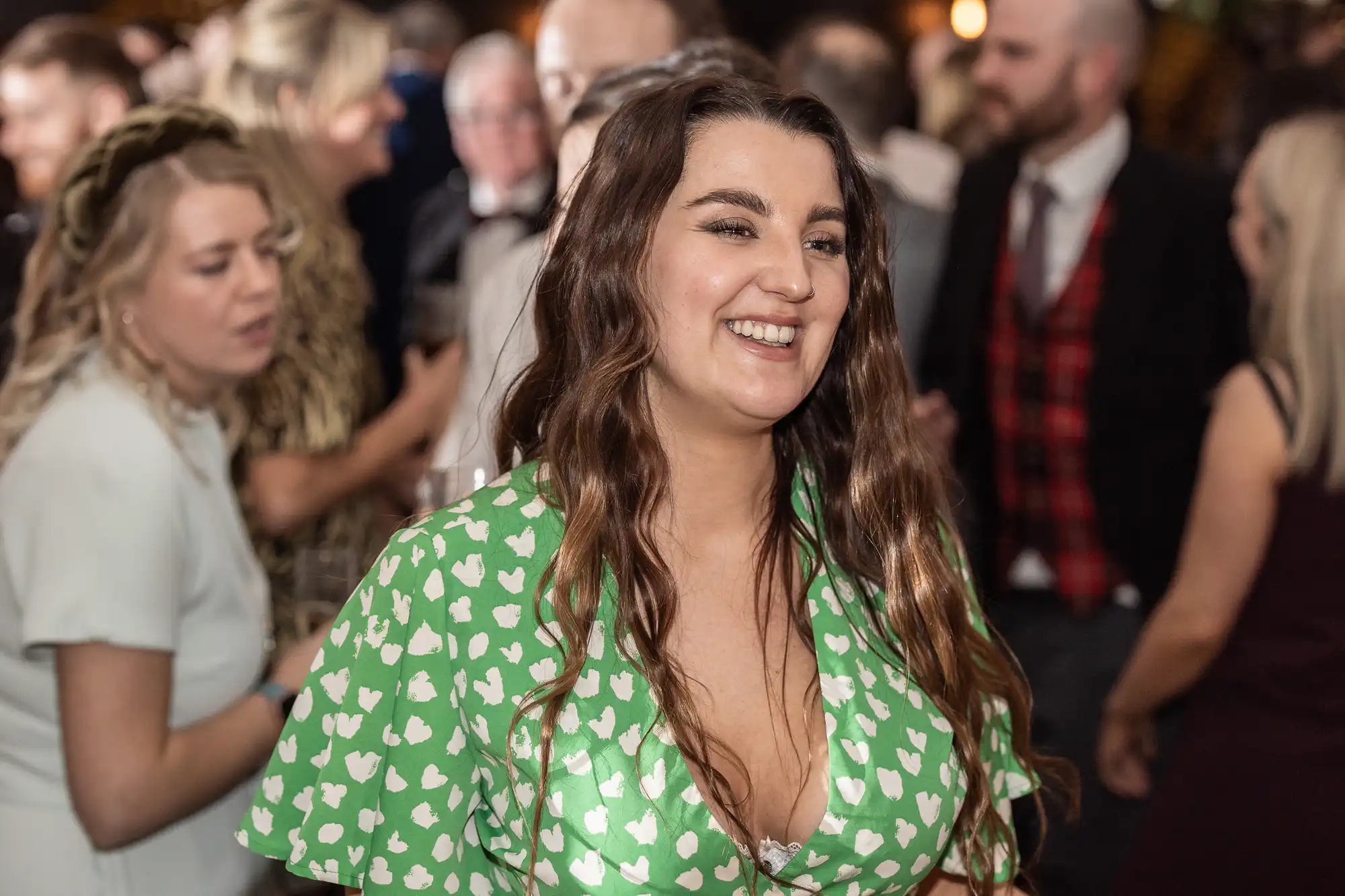 A woman in a green dress with white heart patterns smiles while standing among a group of people at a social event.