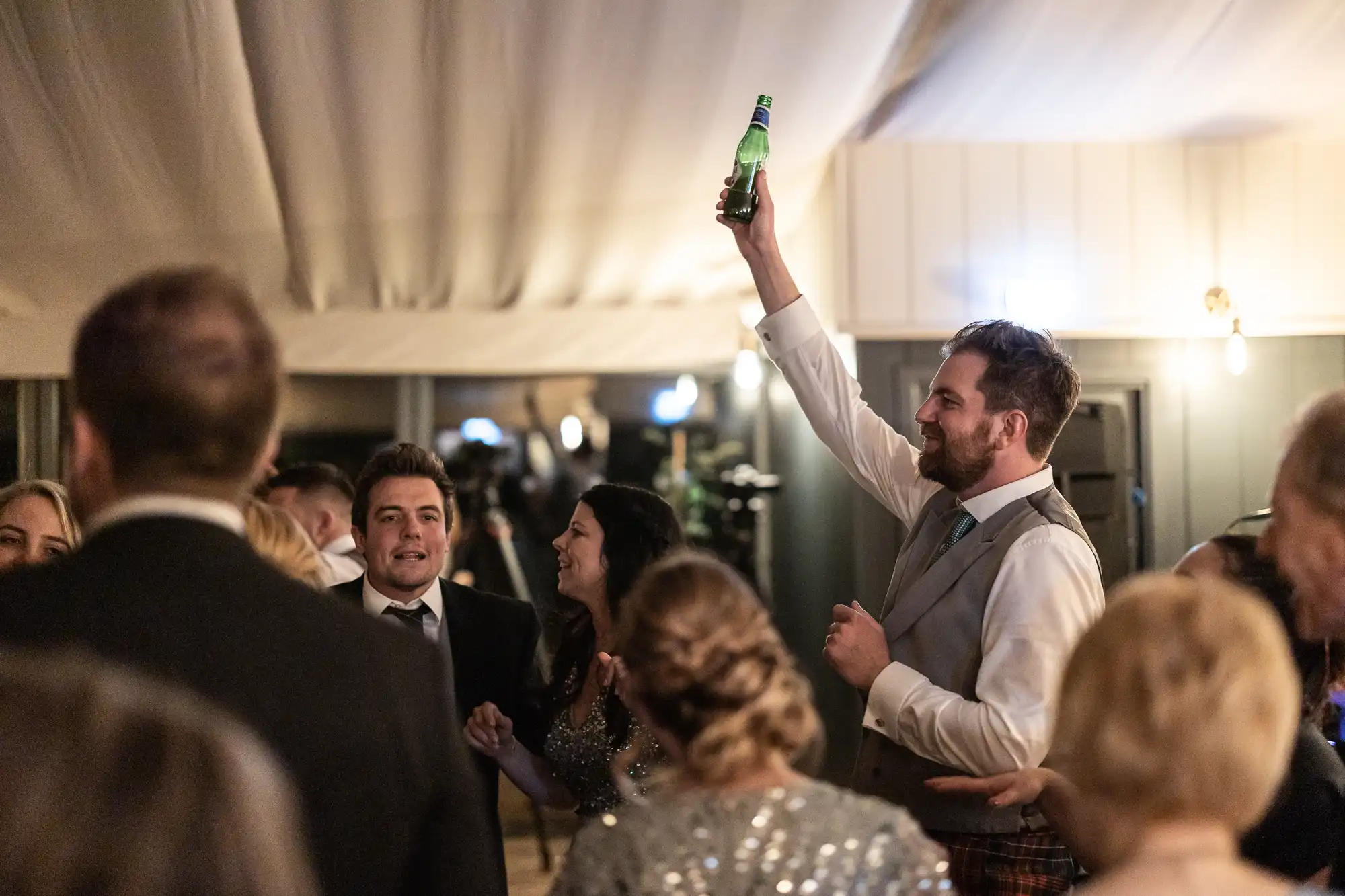 A group of people dressed in formal attire are gathered in a room, with one man raising a green bottle in the air and others around him talking and smiling.