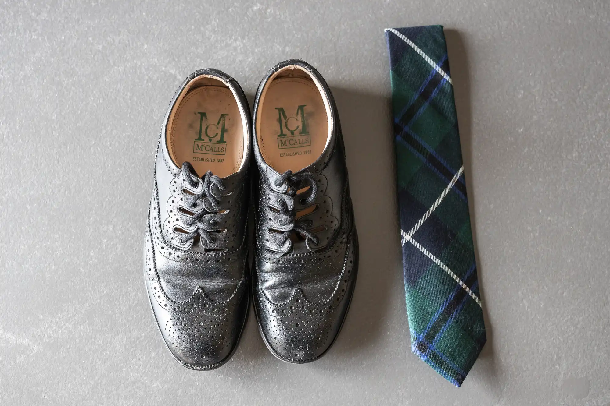 A pair of black dress shoes with brogue detailing placed next to a green and blue plaid necktie on a gray surface.