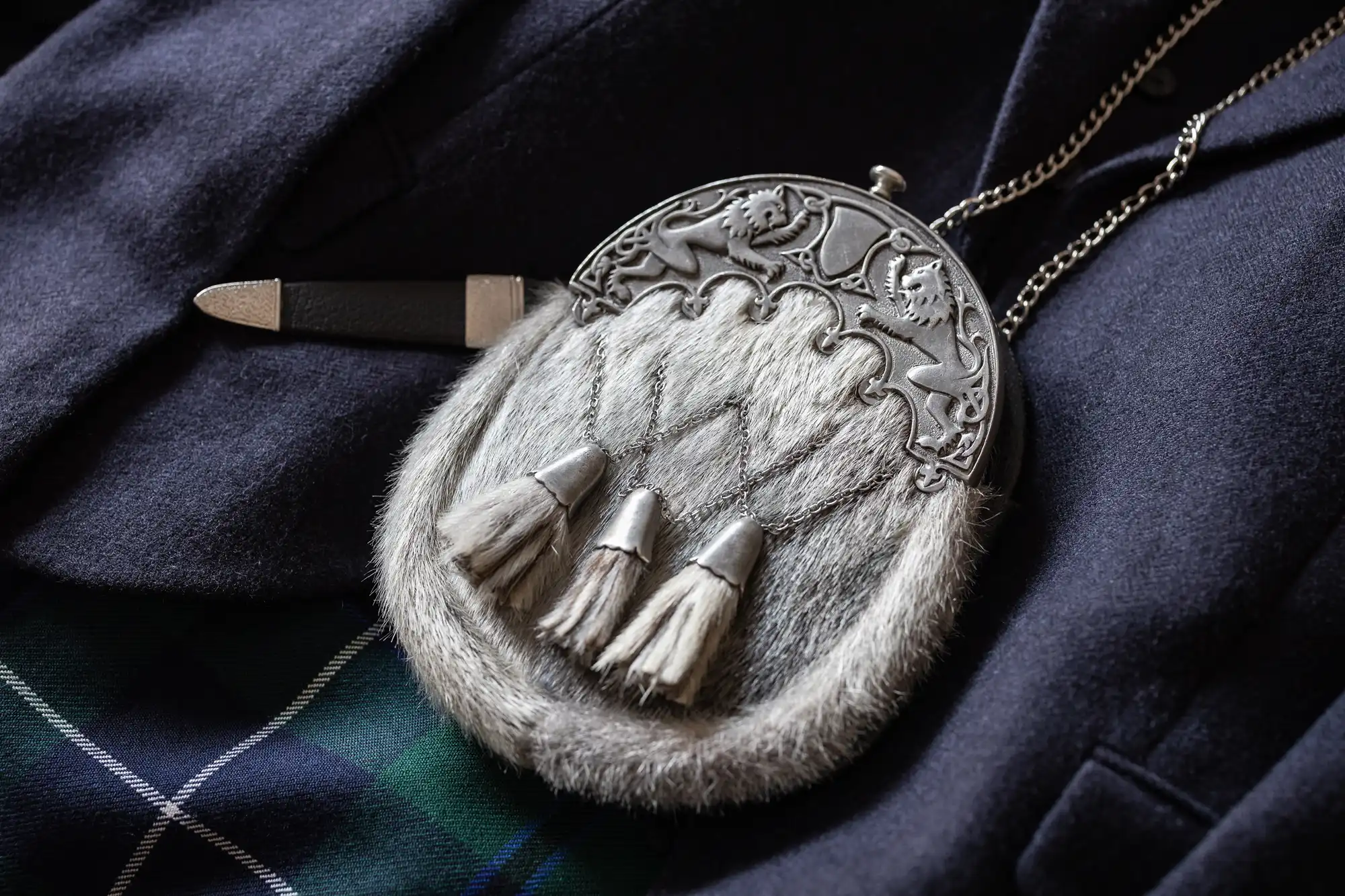 A traditional sporran with metal detailing and tassels rests on dark fabric beside a knife, partially visible against a plaid pattern.