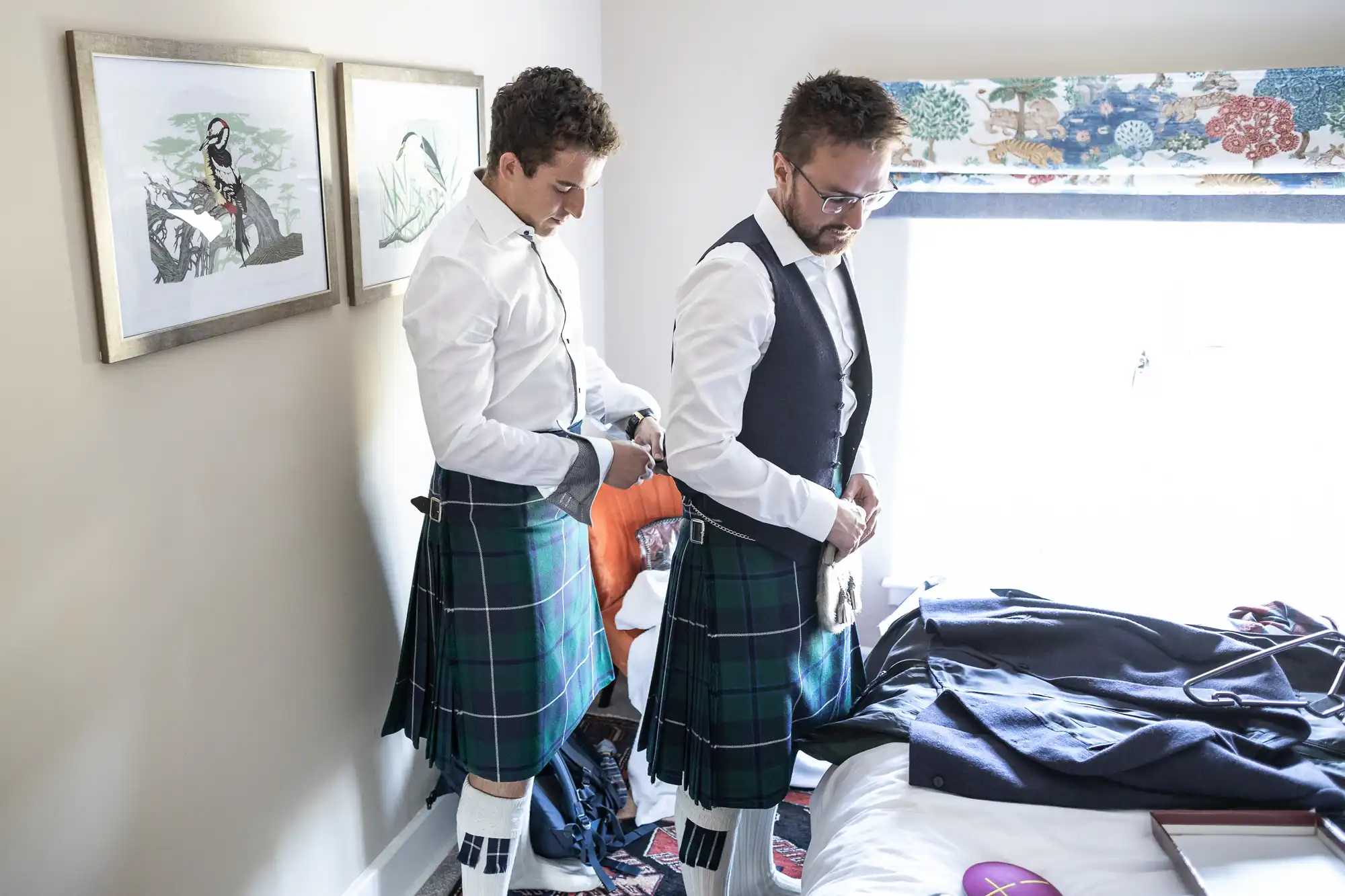 Two men in kilts stand in a room with framed artwork. One man adjusts his clothing while the other appears to be helping. The room has a bed with clothing on it and a window with a patterned blind.