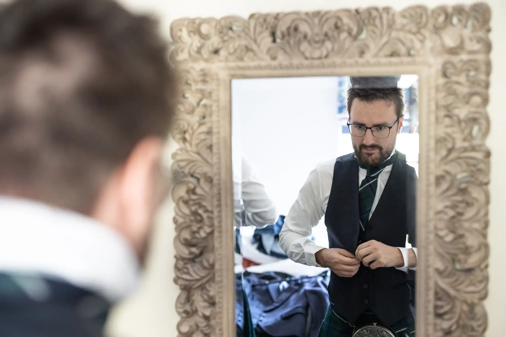 A man wearing glasses and a suit with a kilt looks into a mirror while adjusting his vest.