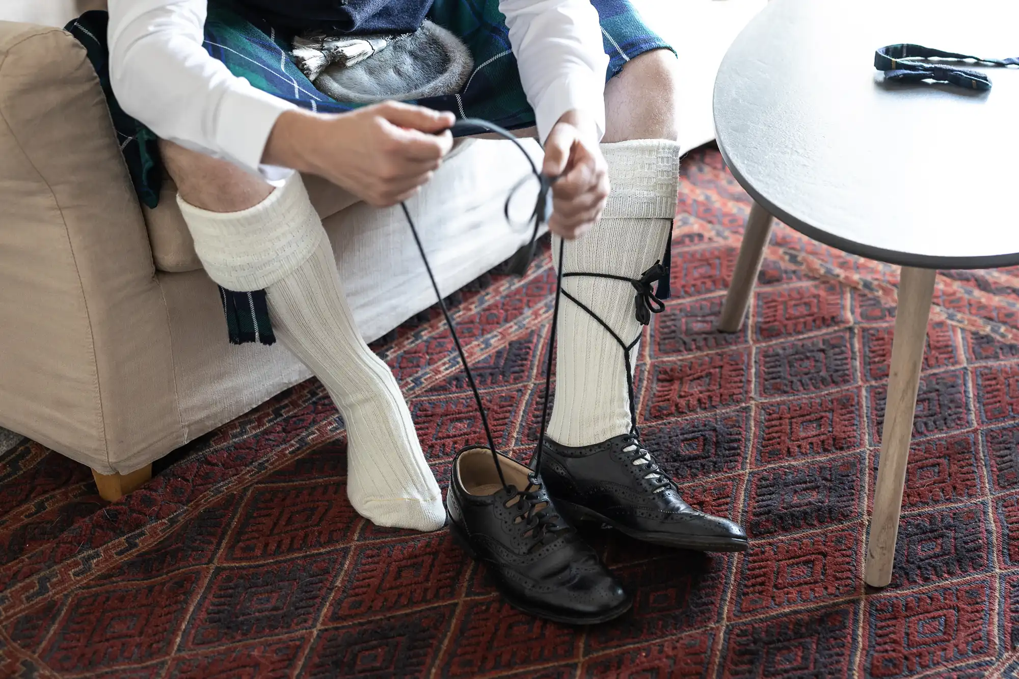 Person sitting on a couch tying black dress shoes while wearing kilt attire, including long white socks and a tartan pattern.