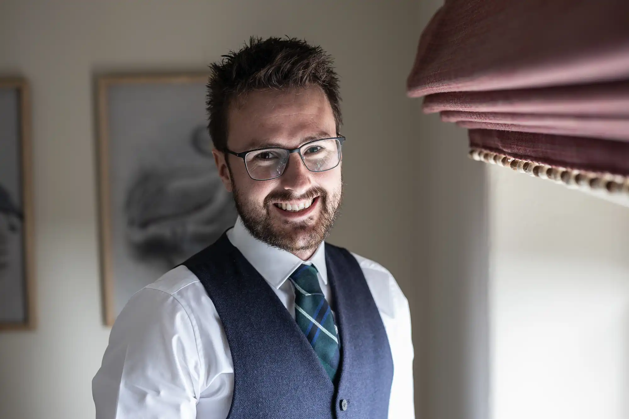 A man wearing glasses, a white shirt, a vest, and a striped tie is smiling while standing near a window with a curtain.