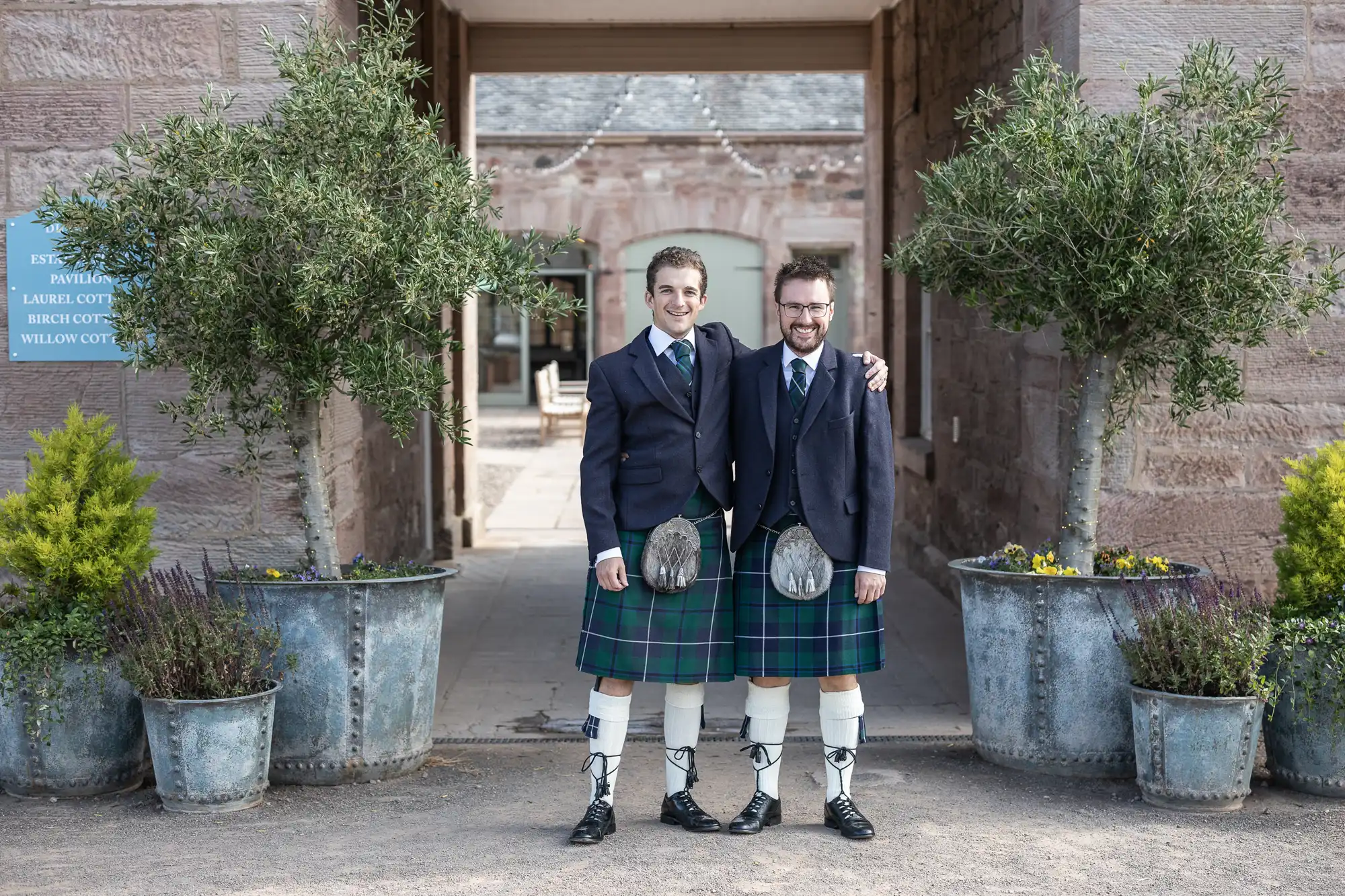 Two people dressed in traditional Scottish kilts and jackets stand arm in arm, smiling, between two large potted plants in an outdoor setting.