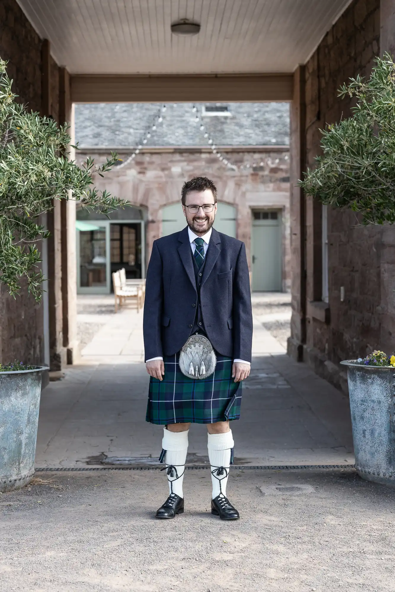 A man smiling, dressed in a traditional Scottish outfit with a kilt and sporran, standing outdoors between two large potted plants.