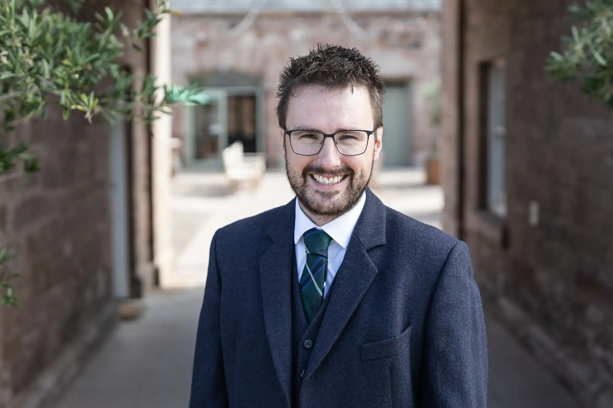 A man with glasses and a beard, dressed in a suit and tie, stands outdoors between brick buildings, smiling at the camera.