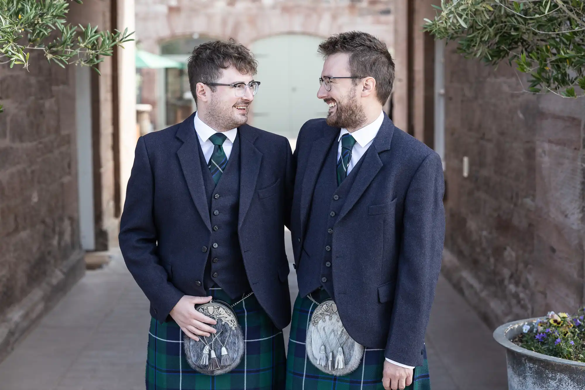 Two men in matching formal attire, including kilts and dark blue jackets, stand in an outdoor walkway, smiling at each other.