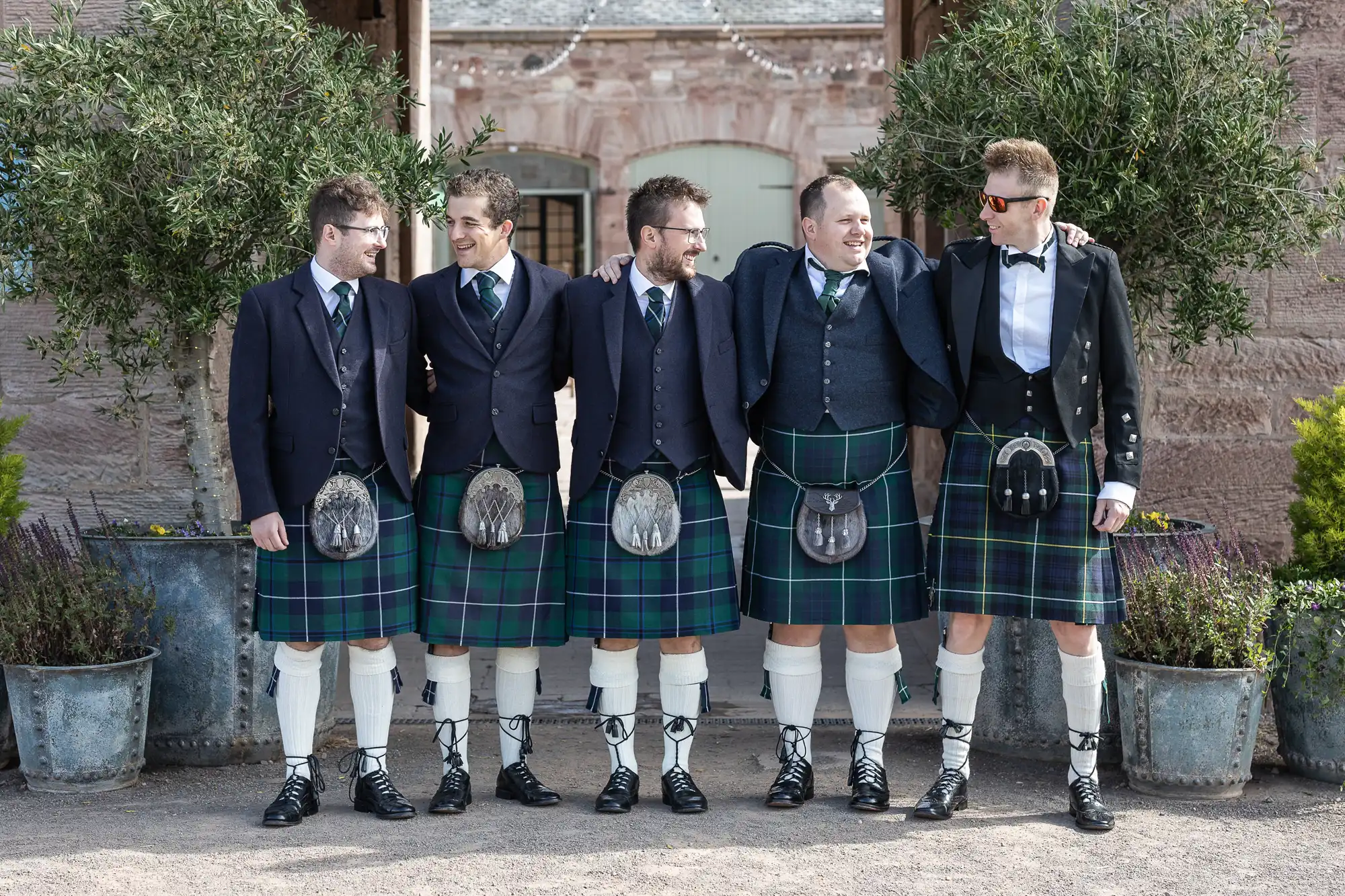 Five men wearing traditional Scottish kilts stand outdoors, arms around each other, smiling. They are dressed in formal attire, including jackets, vests, and ties, with varying styles of kilt accessories.