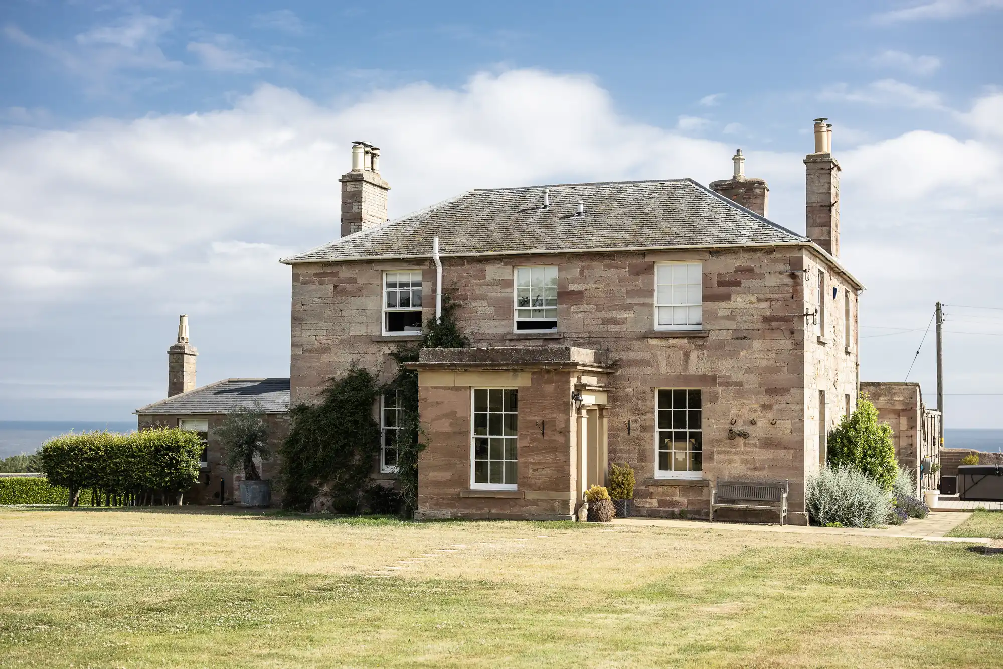 A large, two-story stone house with chimneys, surrounded by a well-maintained lawn, under a blue sky with scattered clouds.