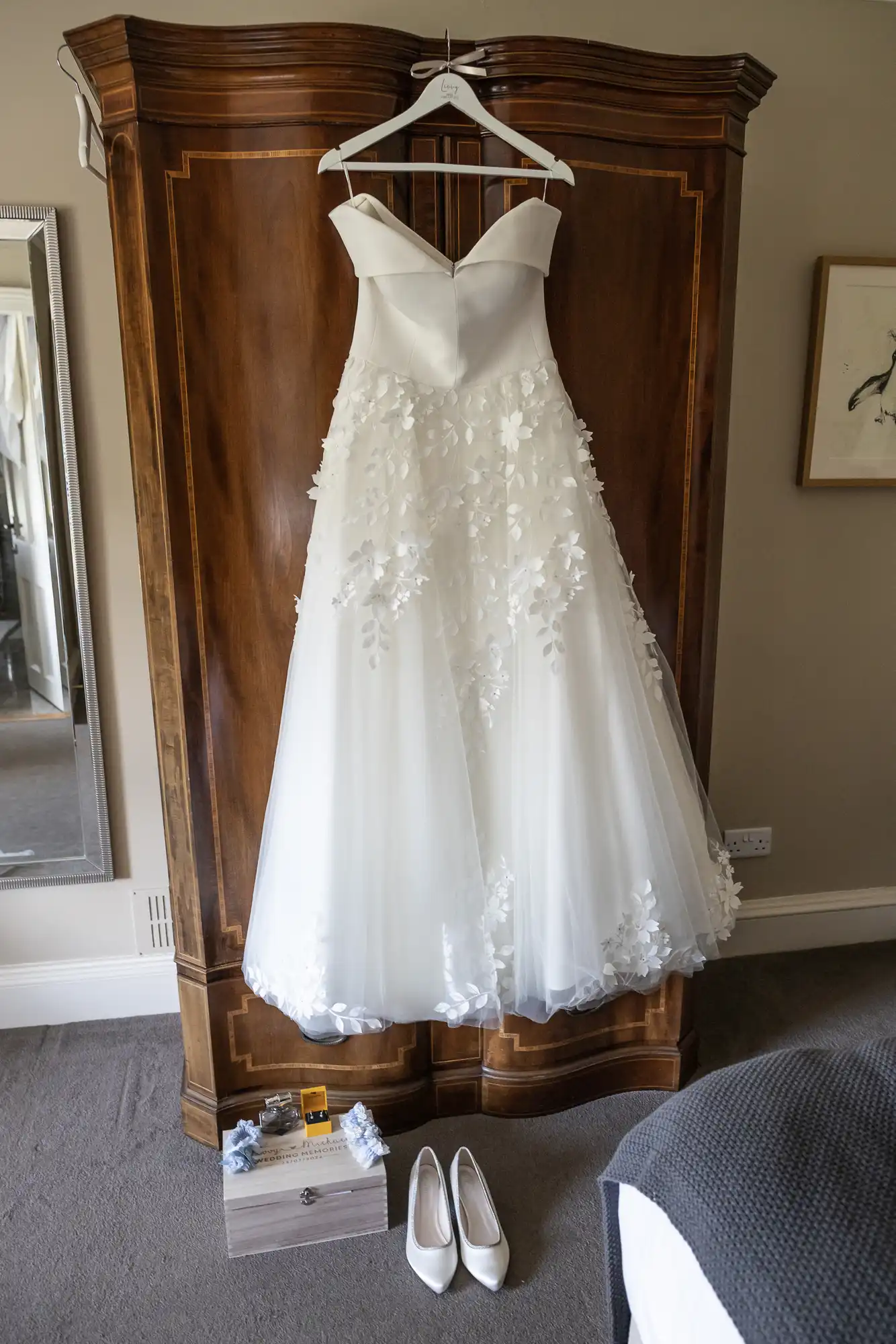 A white strapless wedding dress with floral appliqué detail hangs on a wooden wardrobe. Below are a pair of white shoes, a wooden box, and arranged blue flowers.