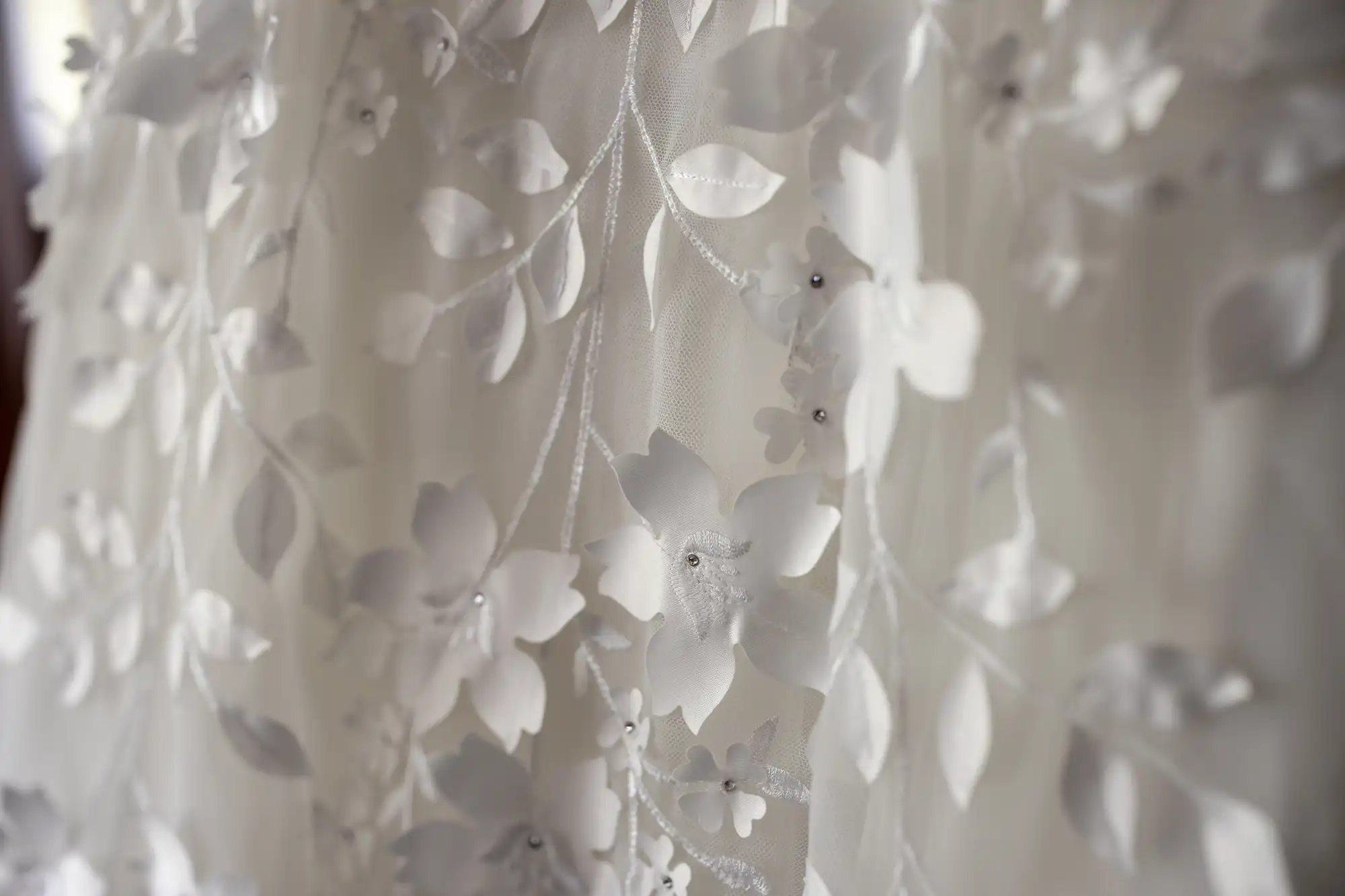 Close-up of white floral embroidery on a sheer fabric. The design features delicate flowers and leaves, creating a subtle and elegant pattern.