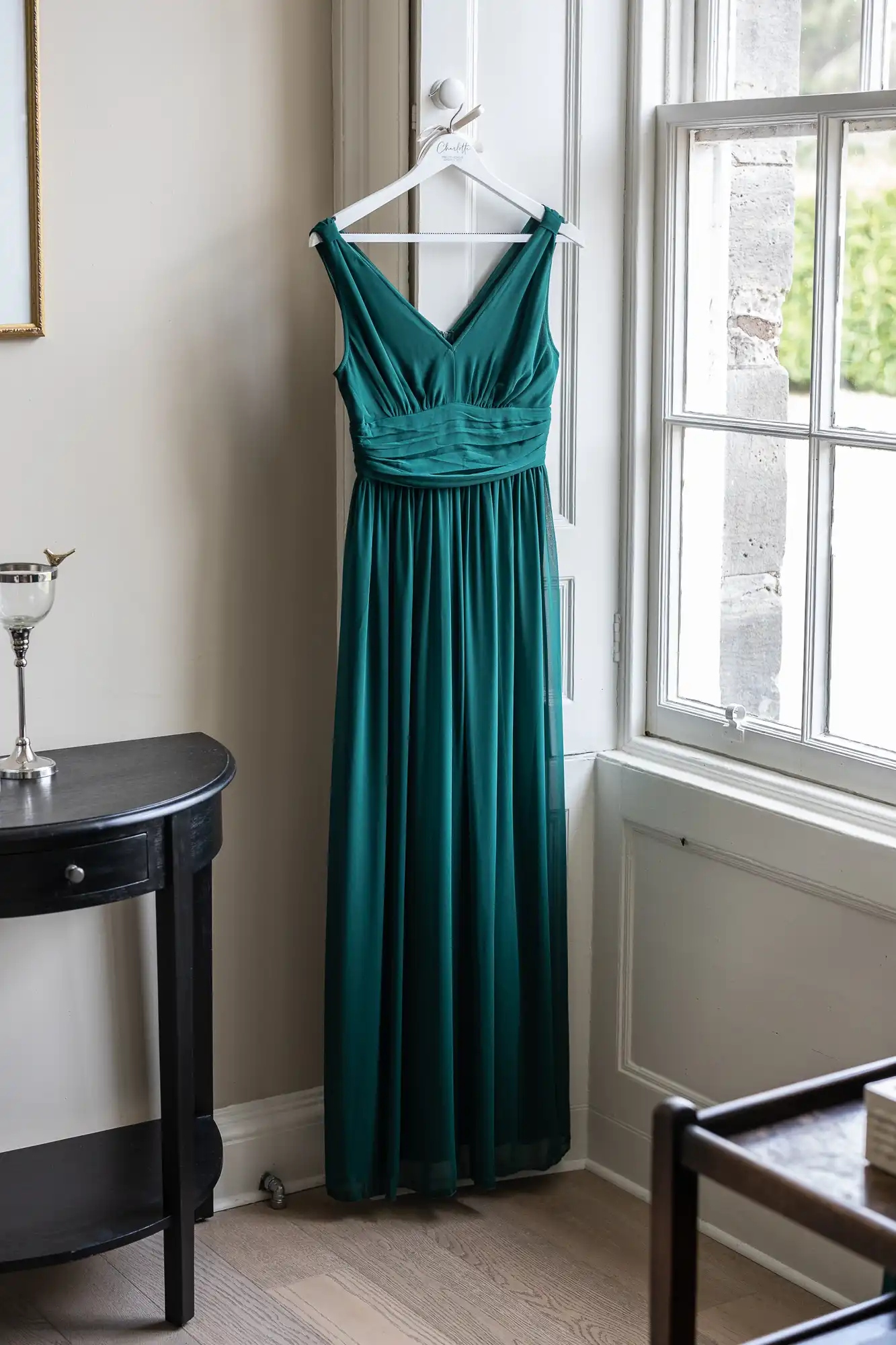 A sleeveless forest green dress hangs on a white hanger next to a window, with a black side table and a goblet placed on it in the foreground.