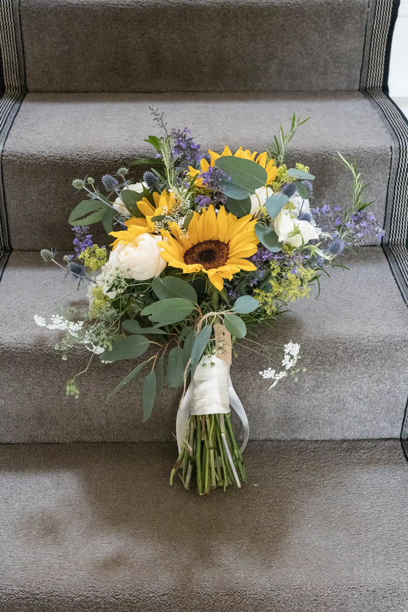 A bouquet of sunflowers, white roses, and greenery is placed on a gray carpeted staircase.