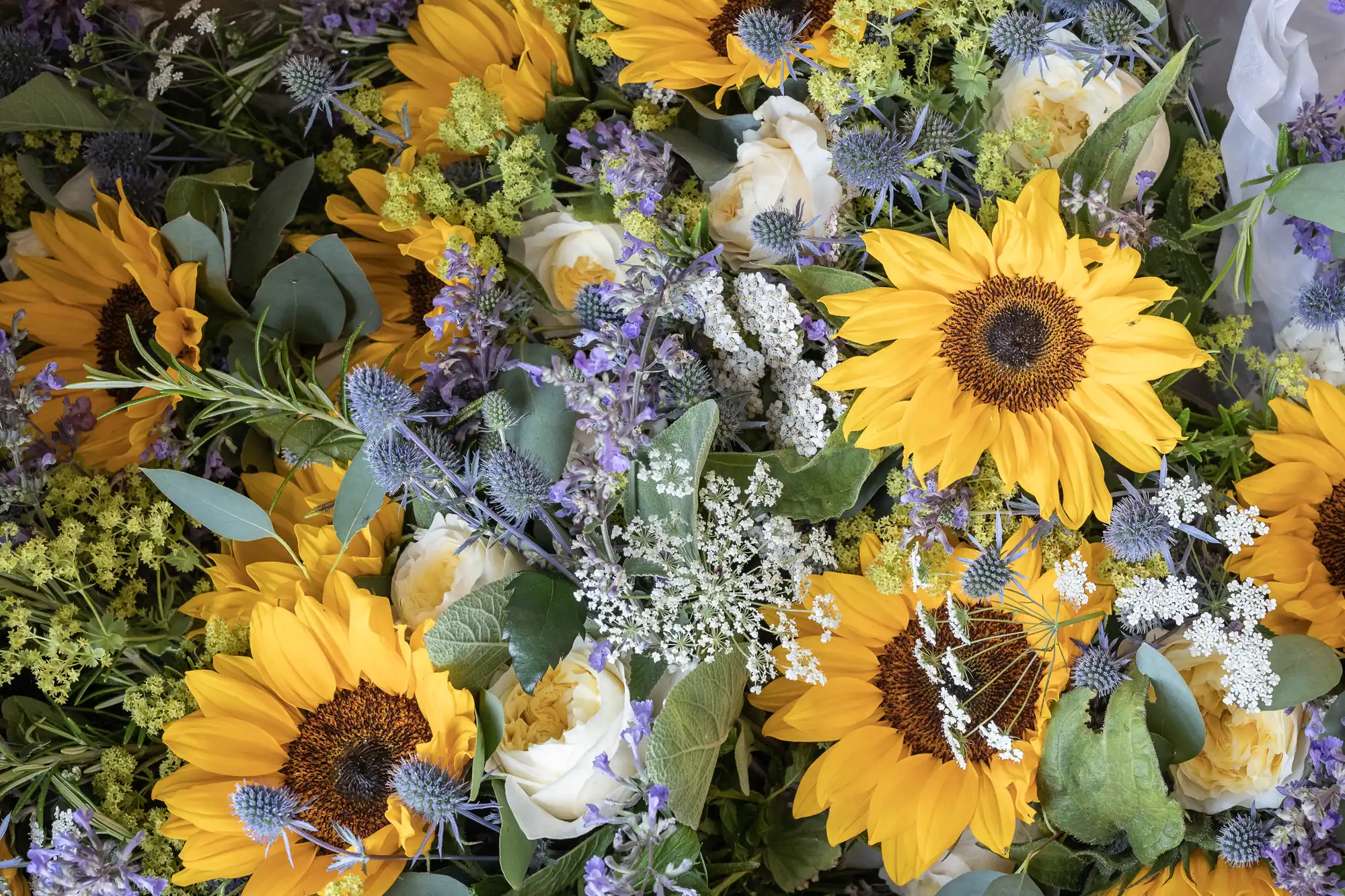 A dense arrangement of yellow sunflowers, white roses, and various green and purple foliage.