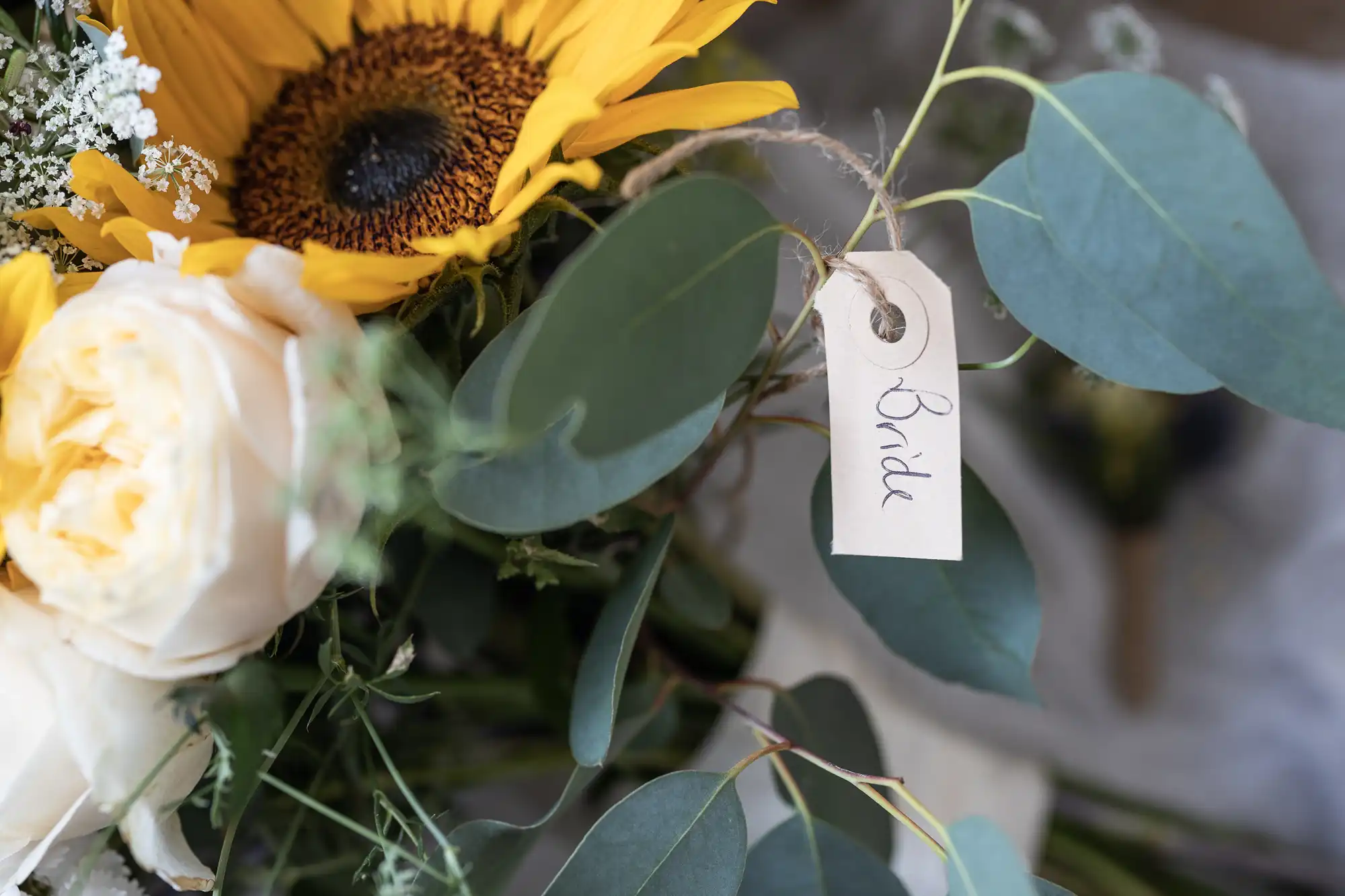 A close-up of a bridal bouquet featuring a sunflower, white roses, and green leaves. A small tag labeled "Bride" is attached to the bouquet.