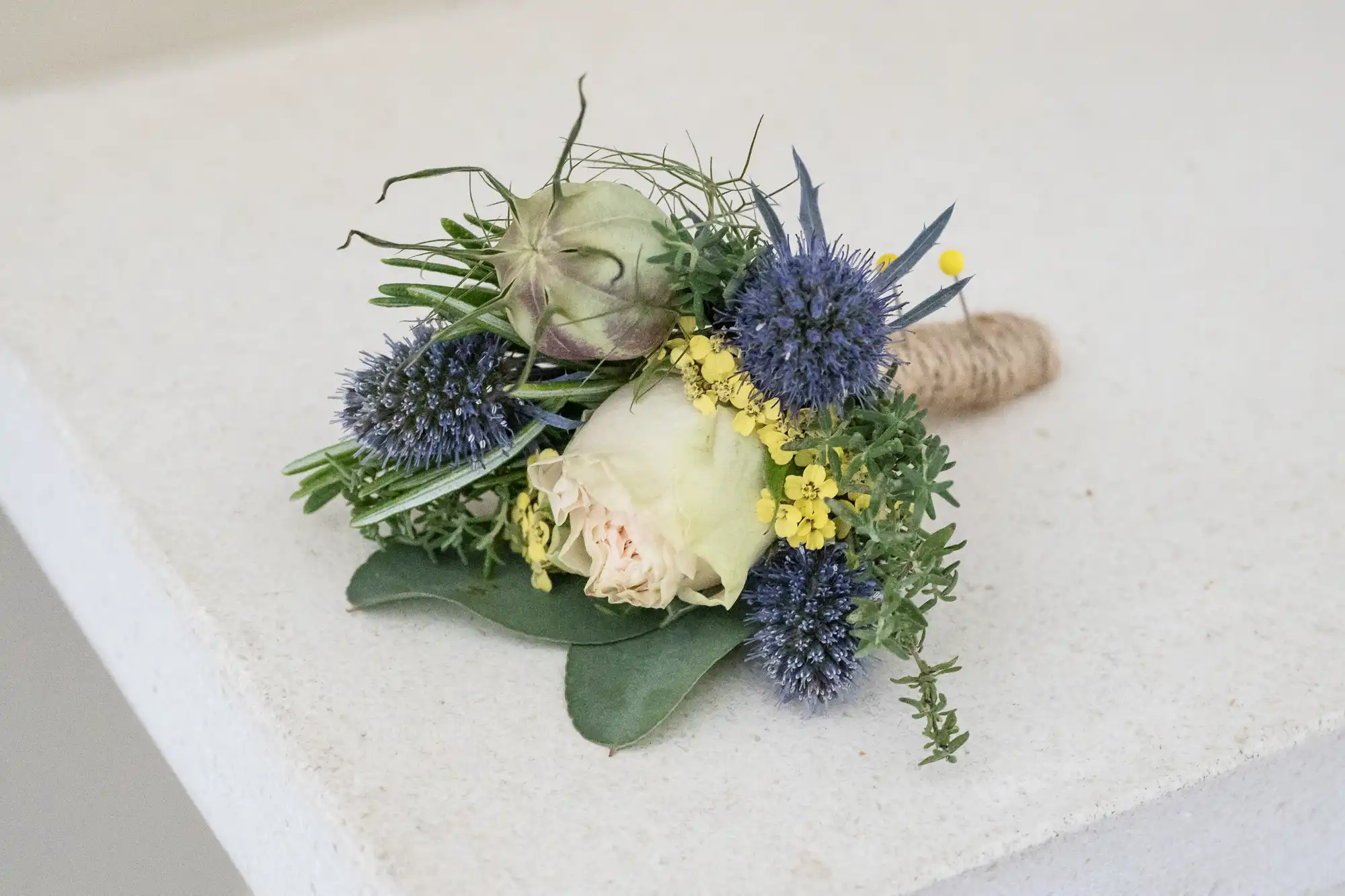 A small floral arrangement with a white rose, blue thistles, yellow flowers, green leaves, and a twine-wrapped stem, placed on a light surface.