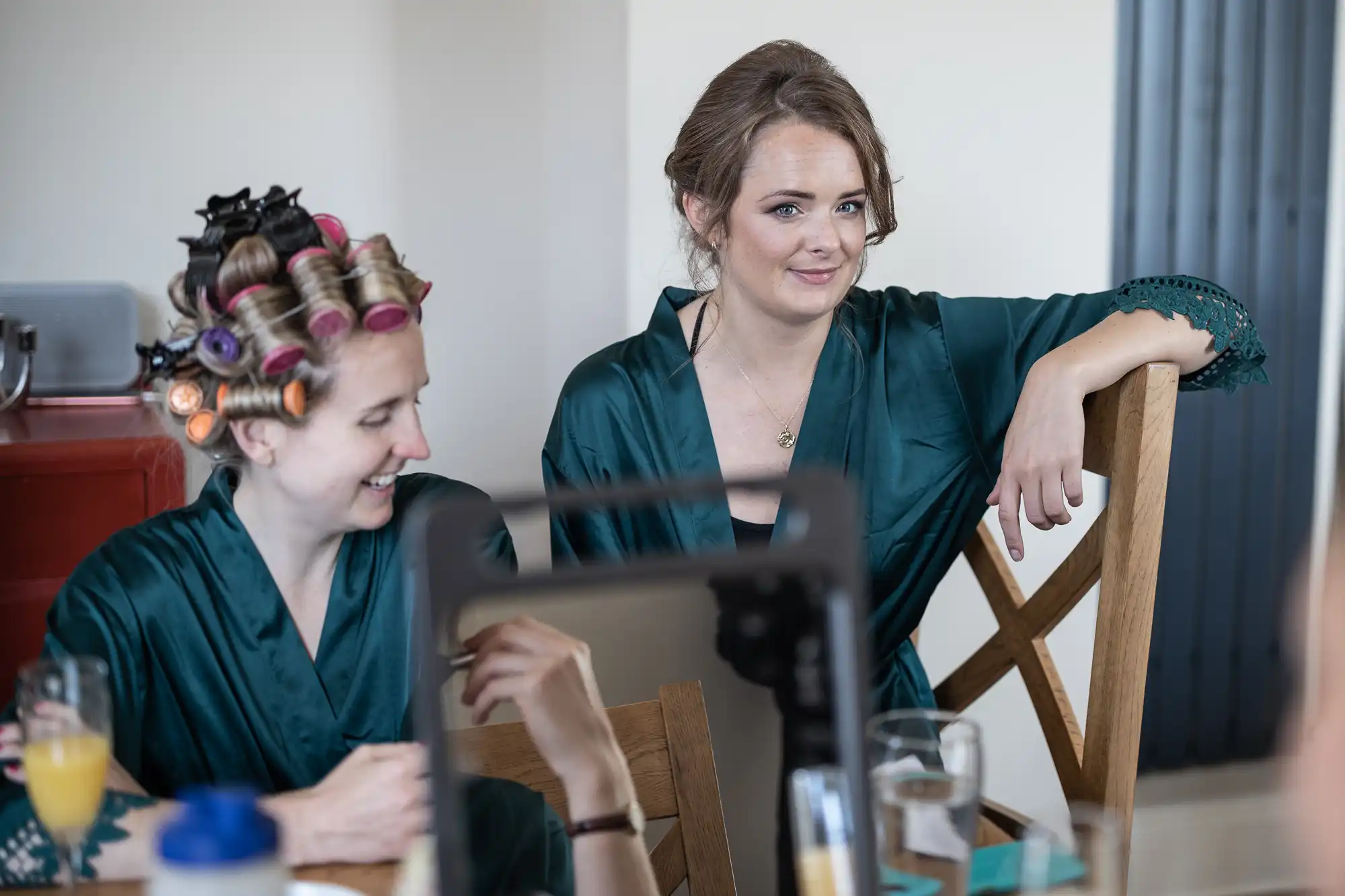 Two women in green robes sit at a table, one with hair rollers and the other leaning on a chair. They appear to be preparing for an event.