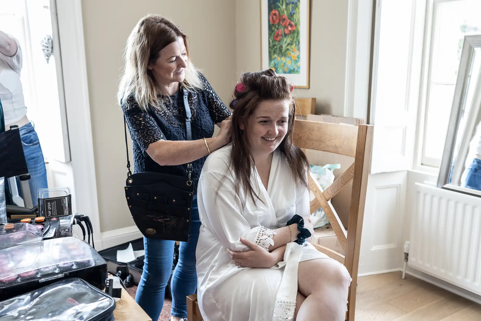 A woman sits on a wooden chair in a white robe while another woman styles her hair using curlers in a well-lit room with a painting on the wall and large windows.